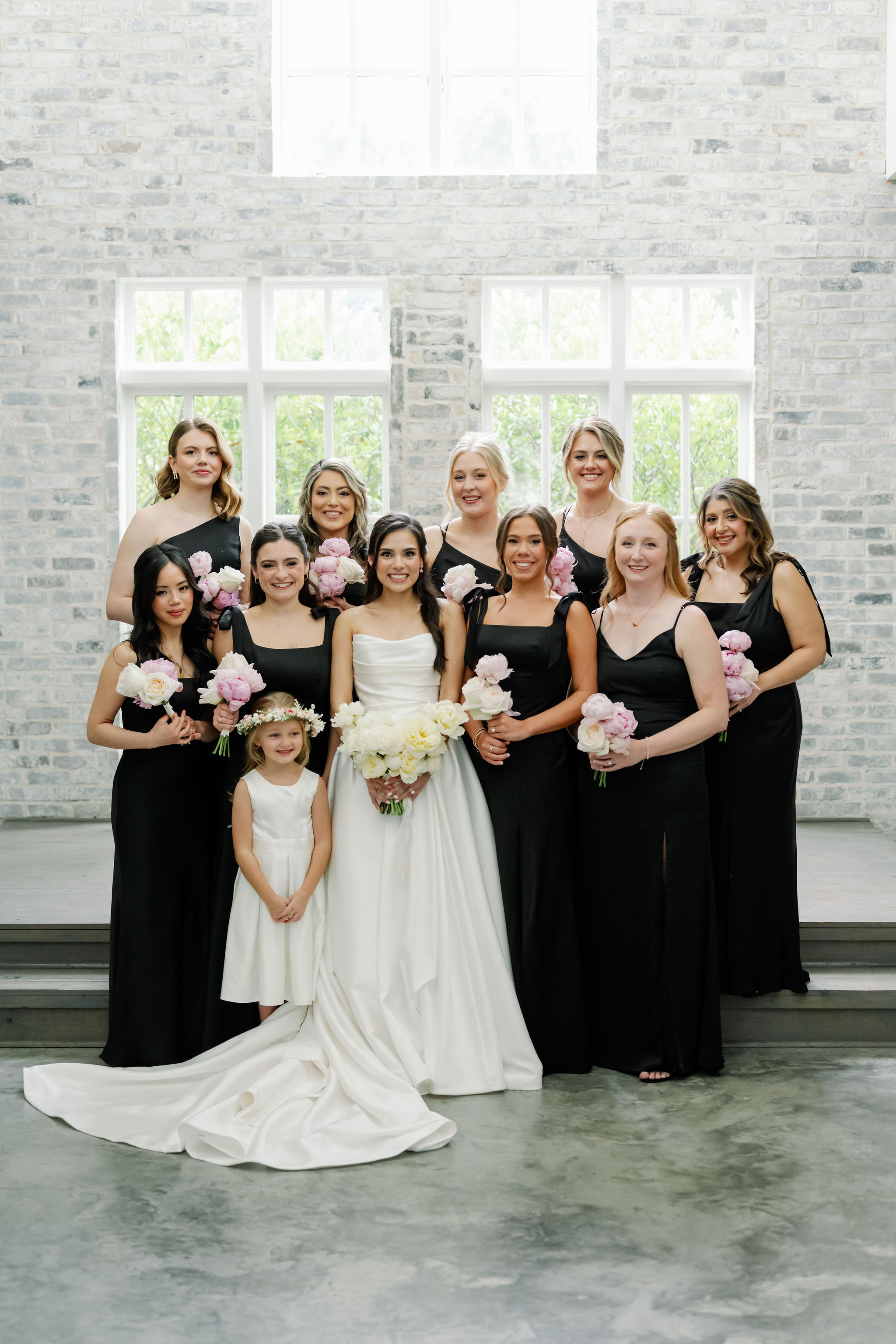 A bride in white surrounded by bridesmaids in black dresses and a flower girl, holding white and soft pink peony bouquets.