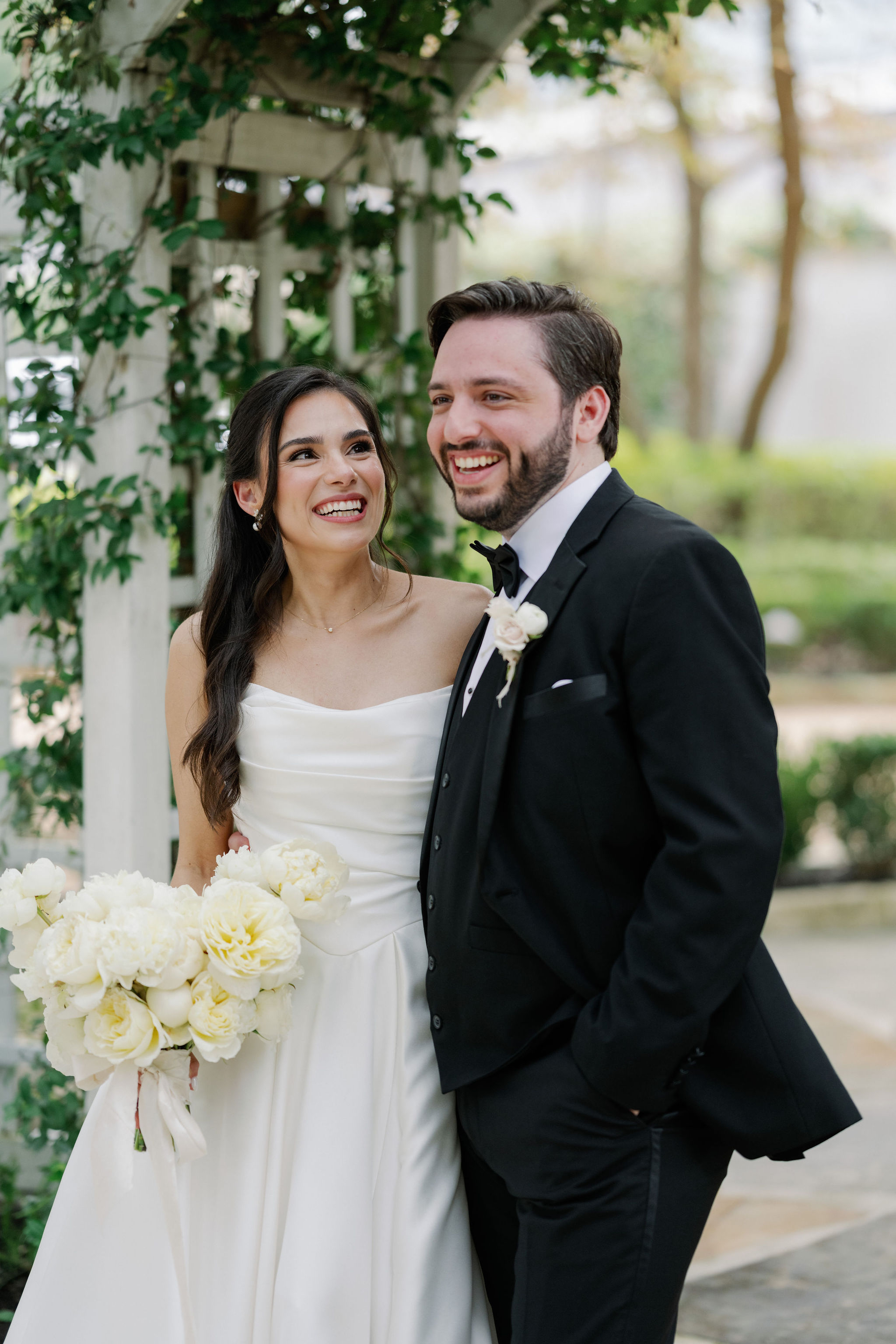 Smiling bride holding an ivory garden rose bouquet standing outdoors with the groom in a black tuxedo.