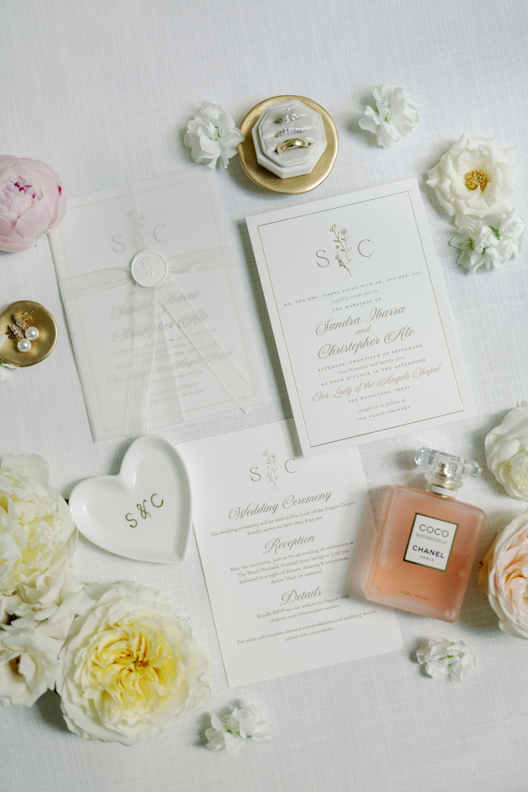 An elegant wedding stationery flat lay with gold-lettered invitations, wedding rings in a velvet box, perfume, and fresh blooms.