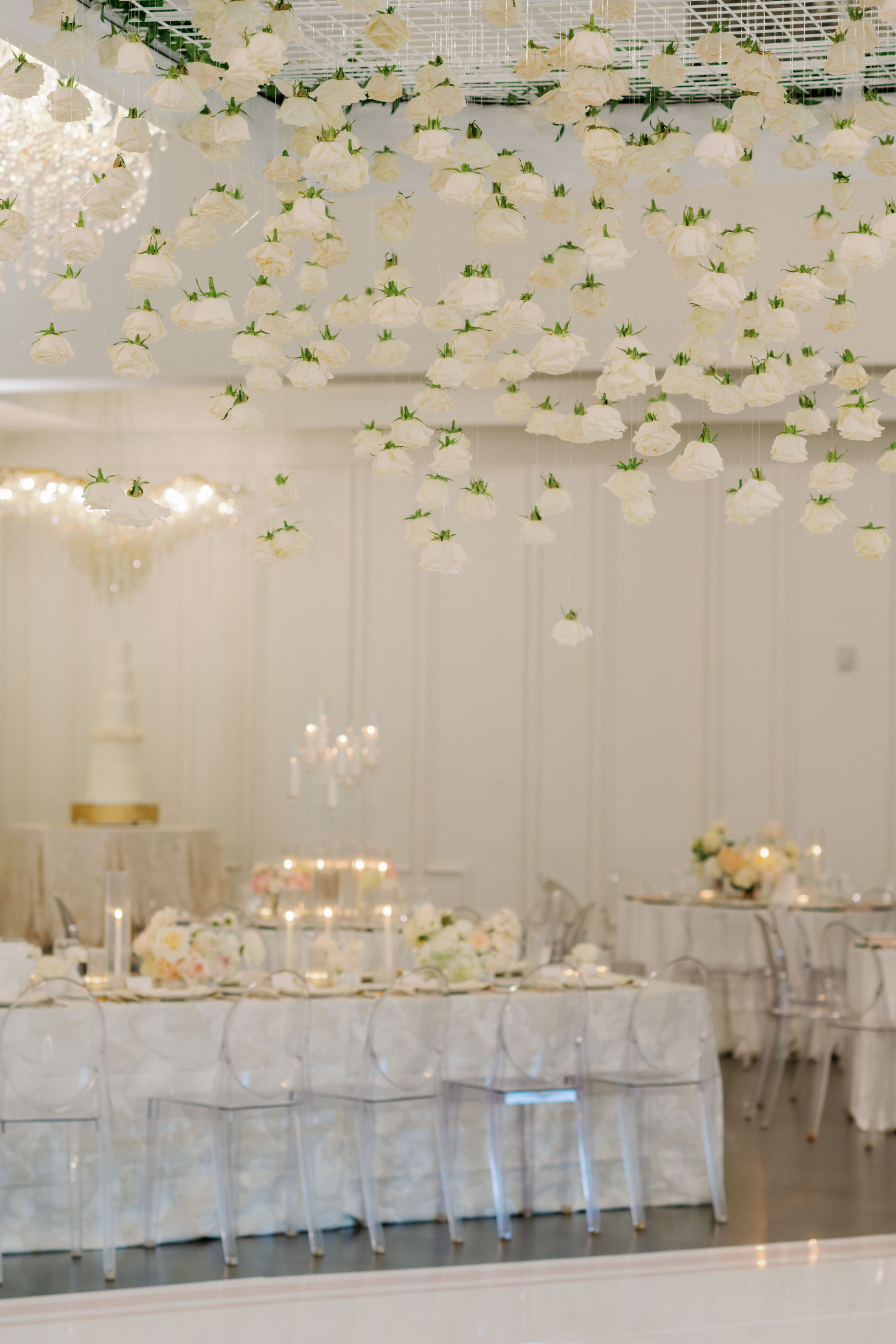 White roses hang suspended from the ceiling above an elegant reception table set with candles and clear chairs.