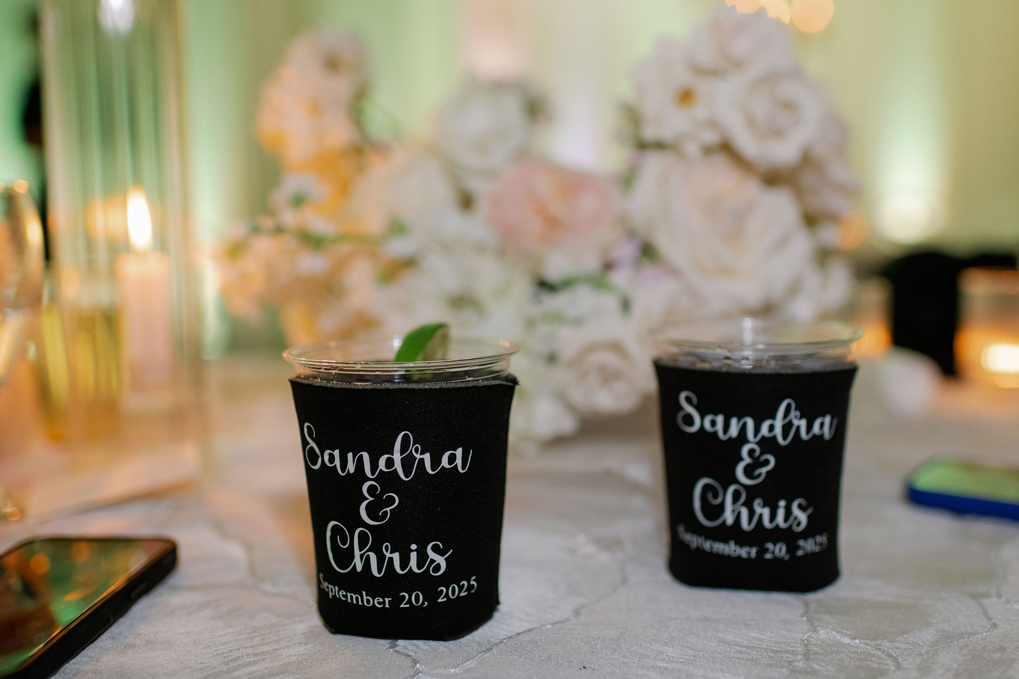 Personalized black cup sleeves holding iced drinks with lime garnish sit before a soft arrangement of white and blush roses.