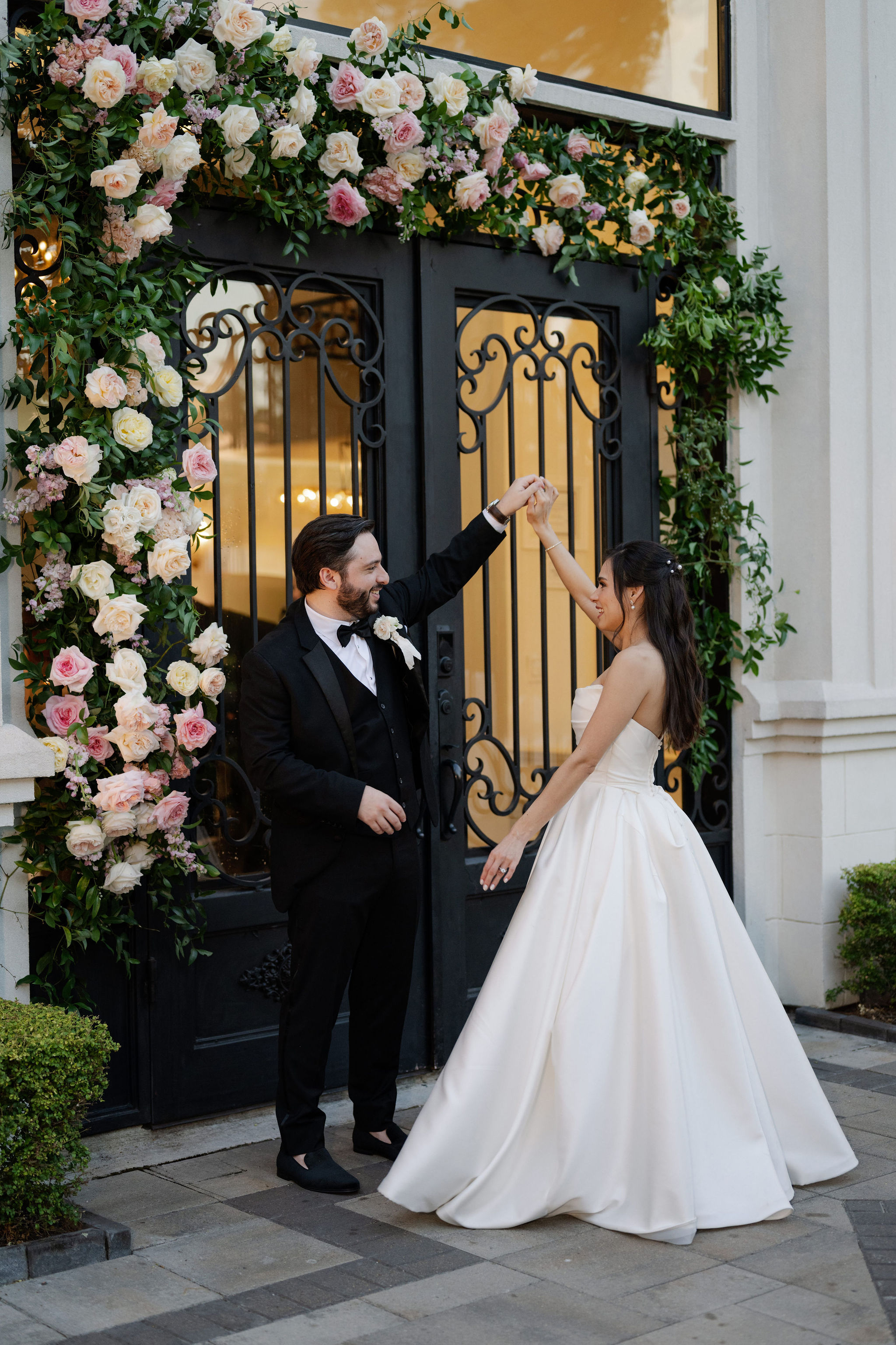 A bride and groom dance before a wrought iron entrance framed by blush, pink, and cream garden roses.