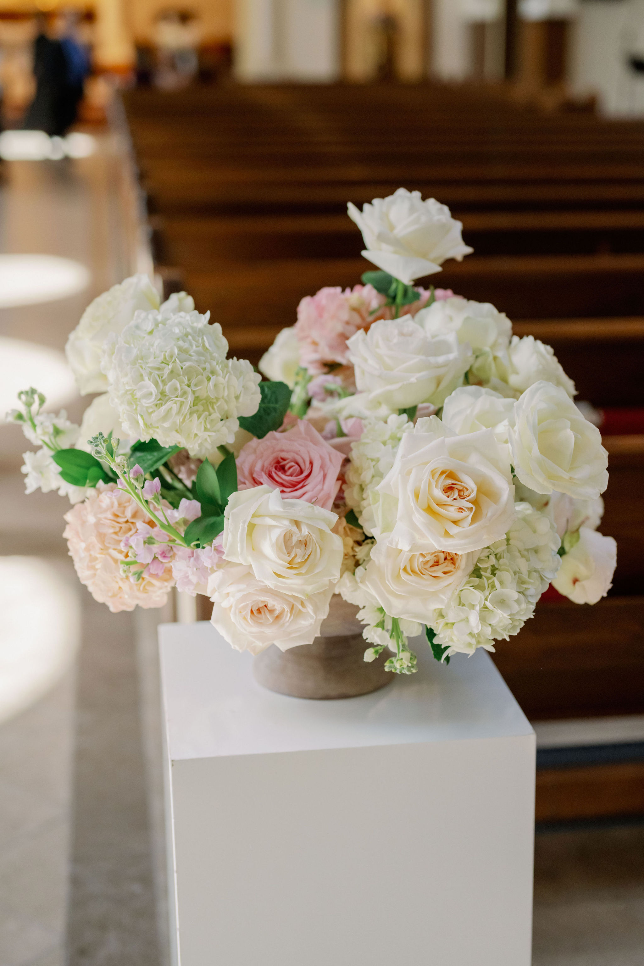 A floral arrangement of white and blush roses and pale hydrangeas resting on a white pedestal.