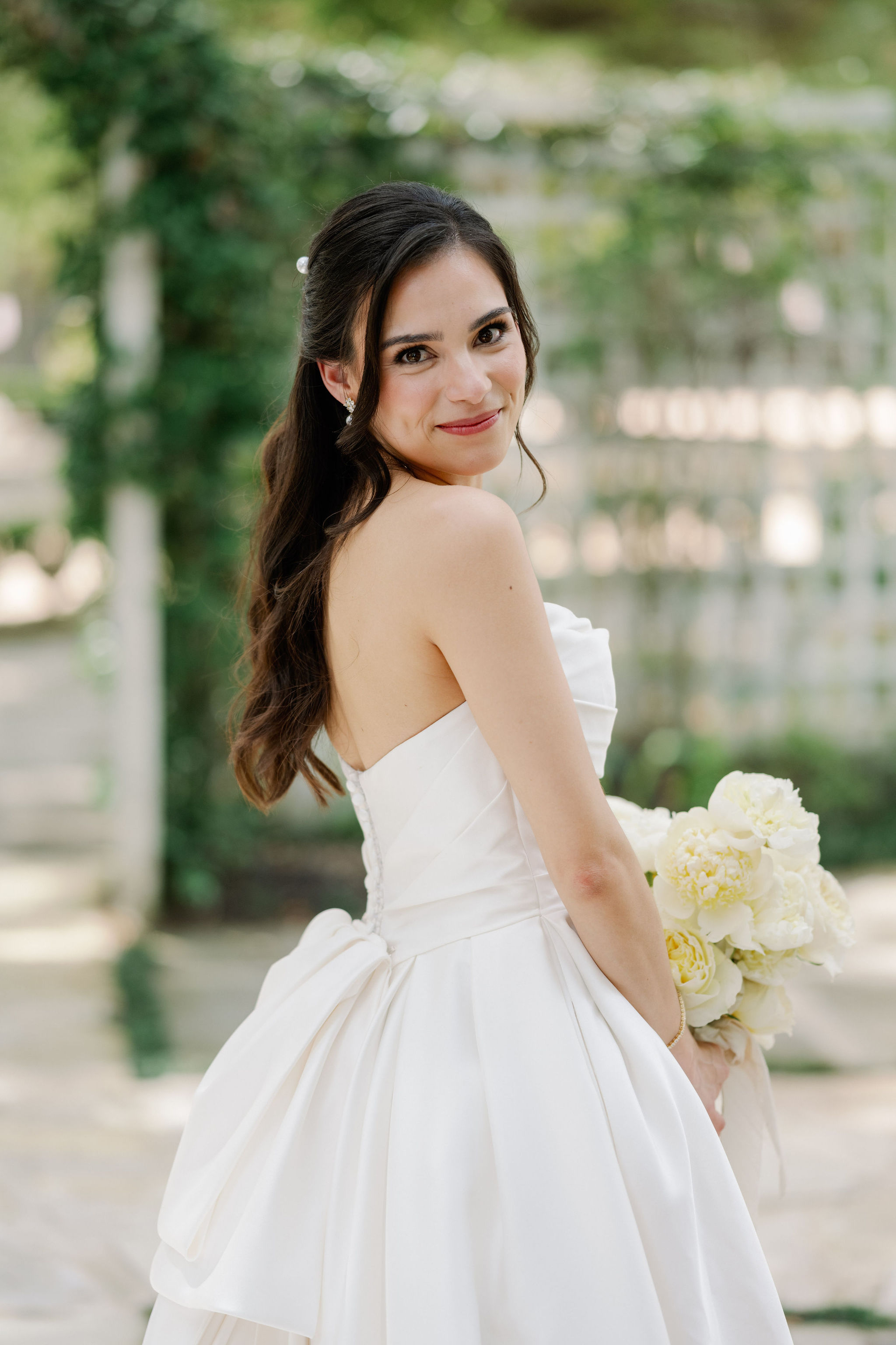 A bride in an elegant strapless satin gown smiles over her shoulder while holding a cream peony bouquet outdoors.