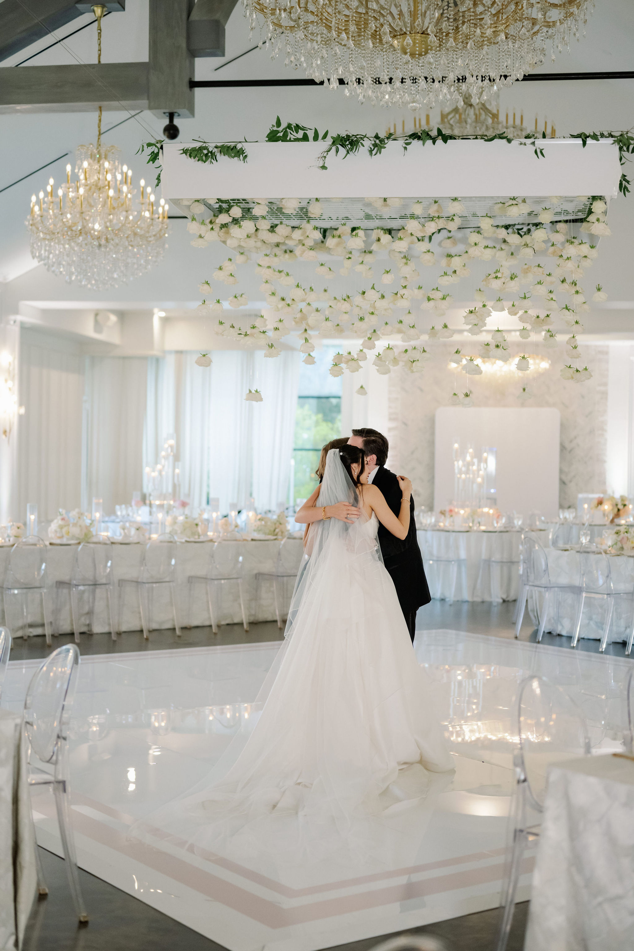 A bride and groom embrace on a glossy white dance floor beneath a suspended canopy of delicate hanging white roses.