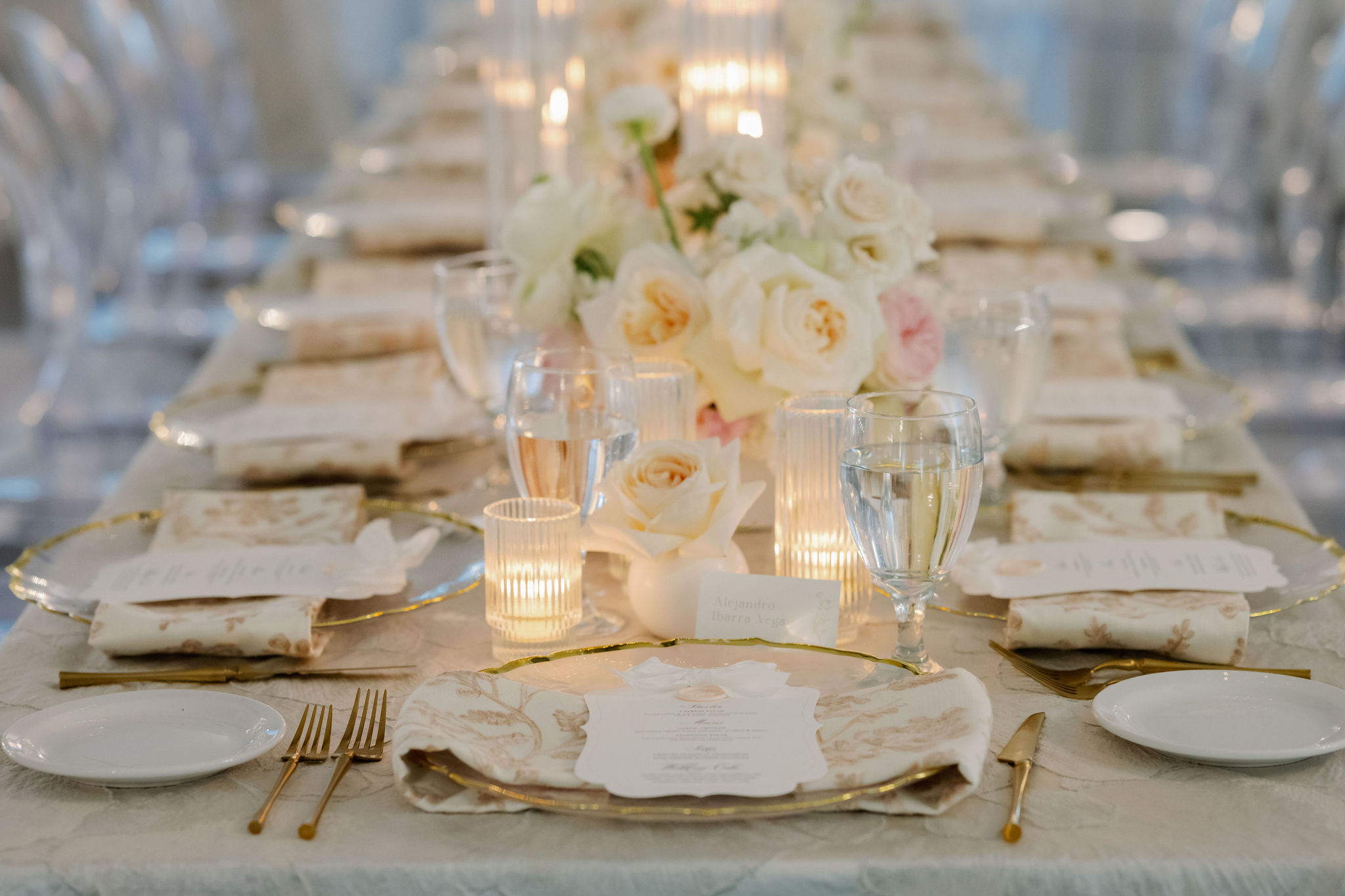 Elegant wedding table setting with cream roses, glowing ribbed candleholders, gold flatware, and patterned napkins.