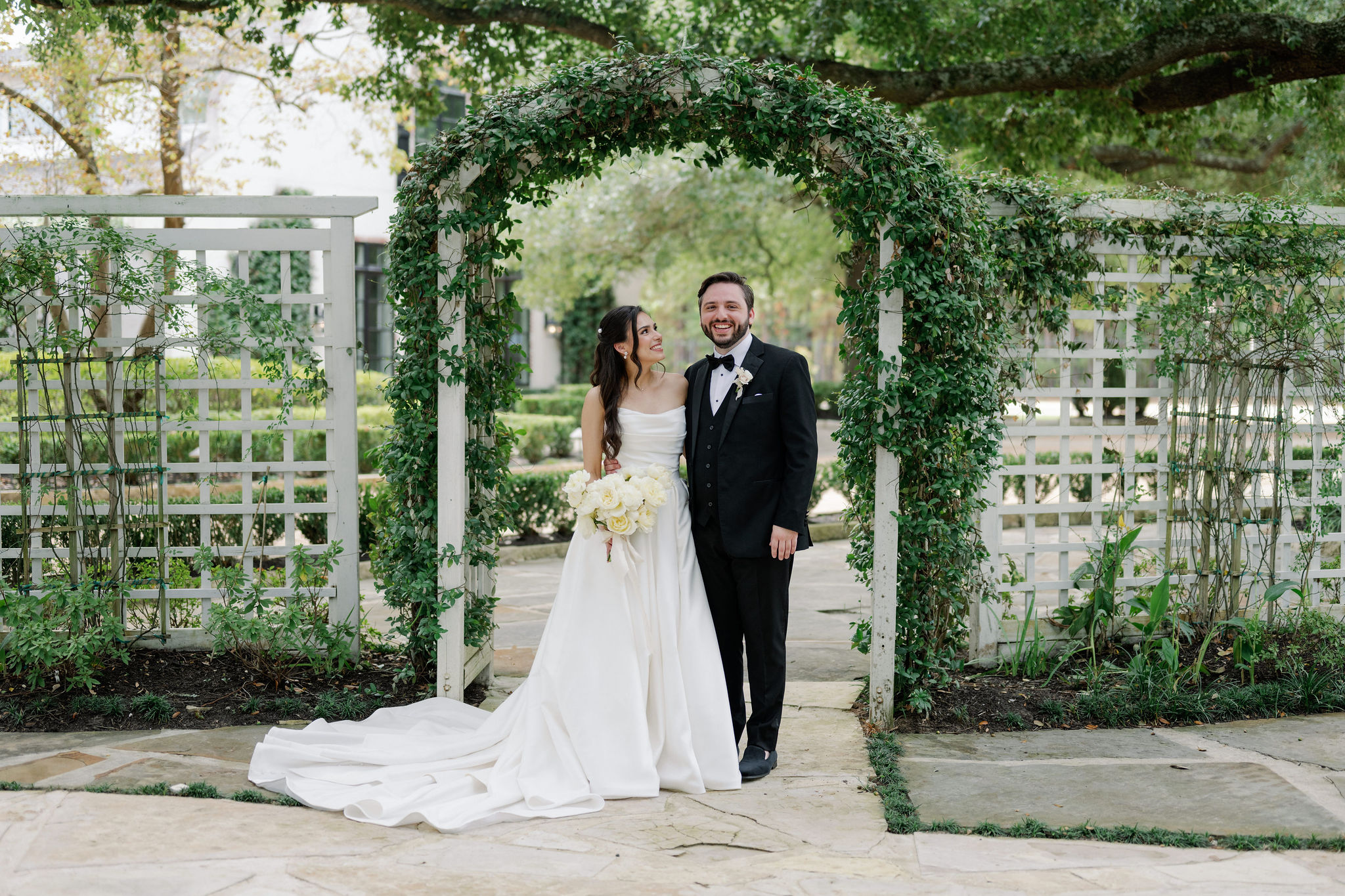 A bride and groom pose together under an ivy-covered garden arbor holding a bouquet of ivory roses.