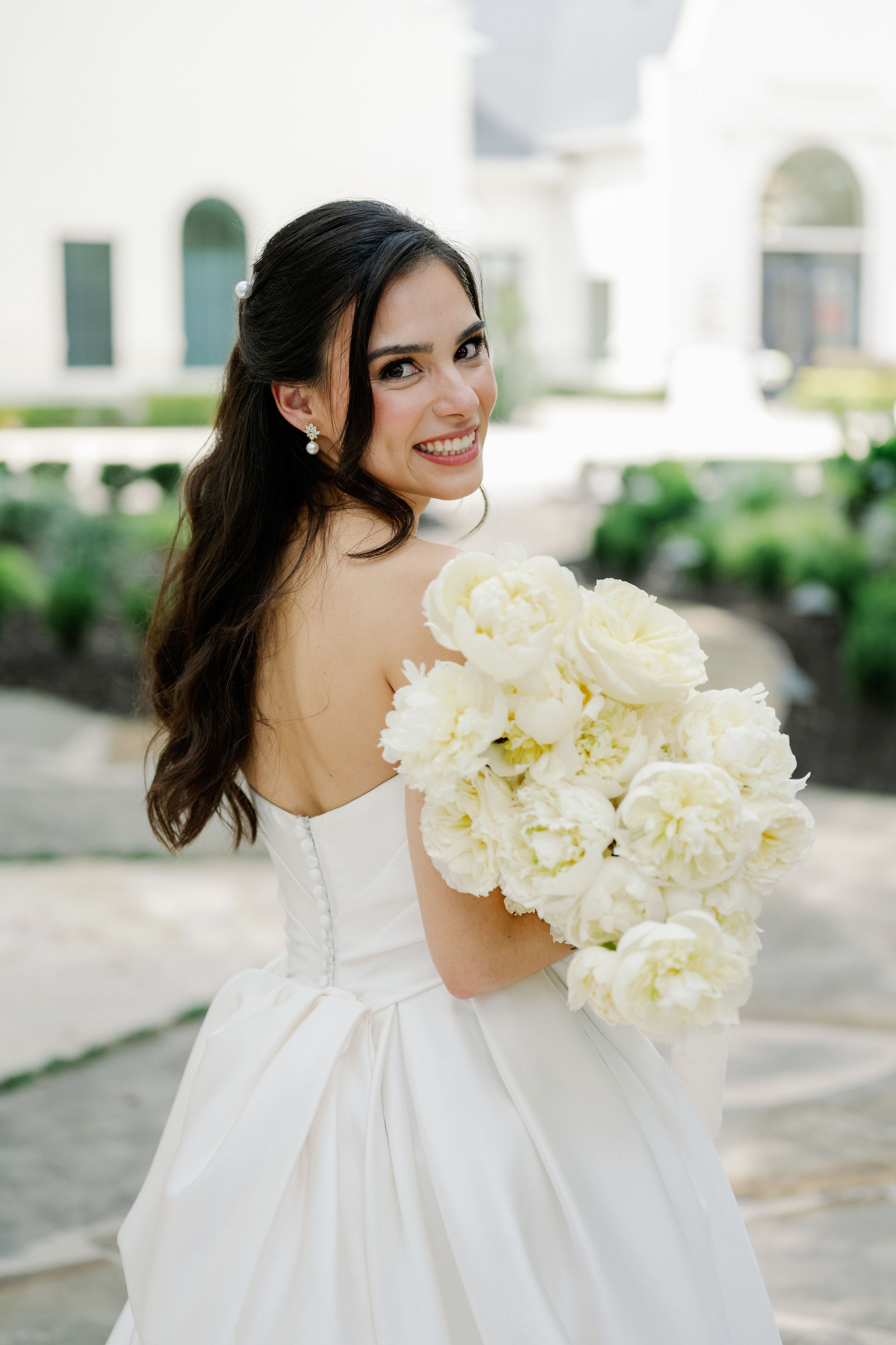A smiling bride in an ivory strapless gown holds a lush bouquet of creamy white peonies and garden roses.