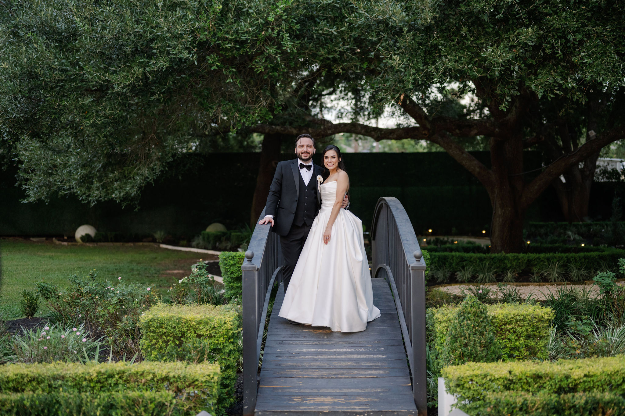 A bride in a satin ballgown and a groom in a black tuxedo pose together on a wooden footbridge in a lush green garden.