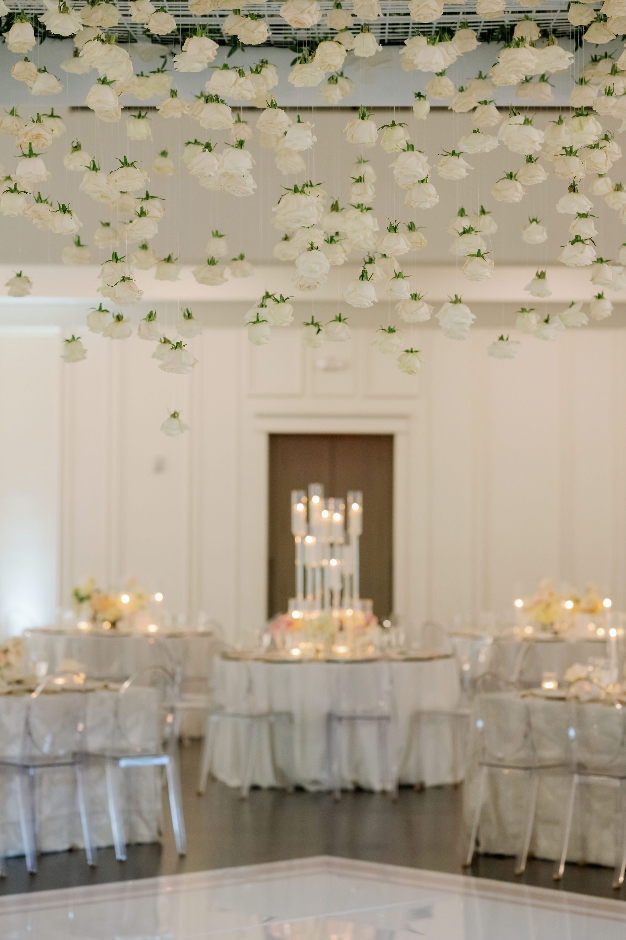 White roses hang suspended from the ceiling above an elegant wedding reception room illuminated by candlelight centerpieces.