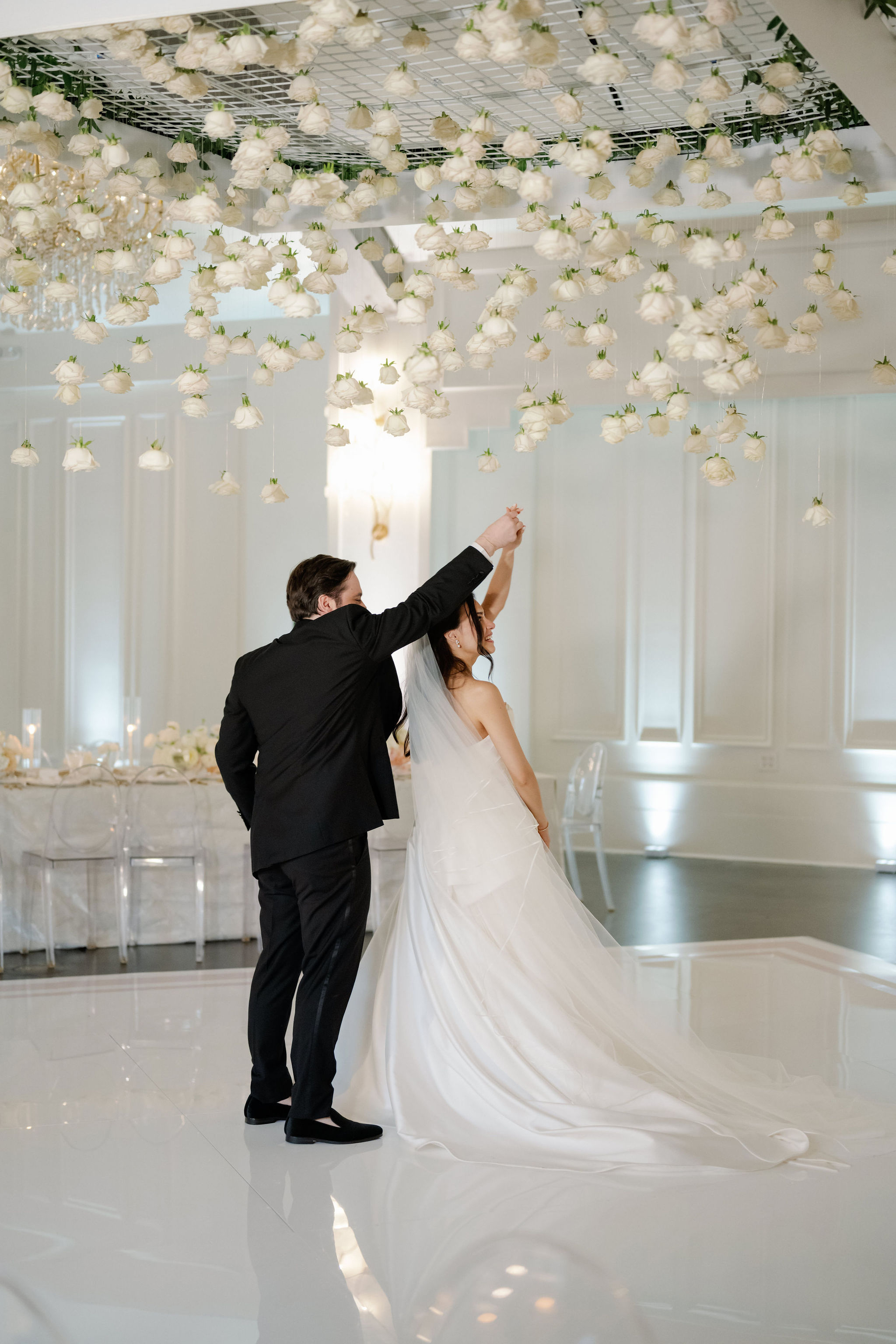 A bride and groom dancing beneath a suspended ceiling installation of hanging white roses on an all-white dance floor.