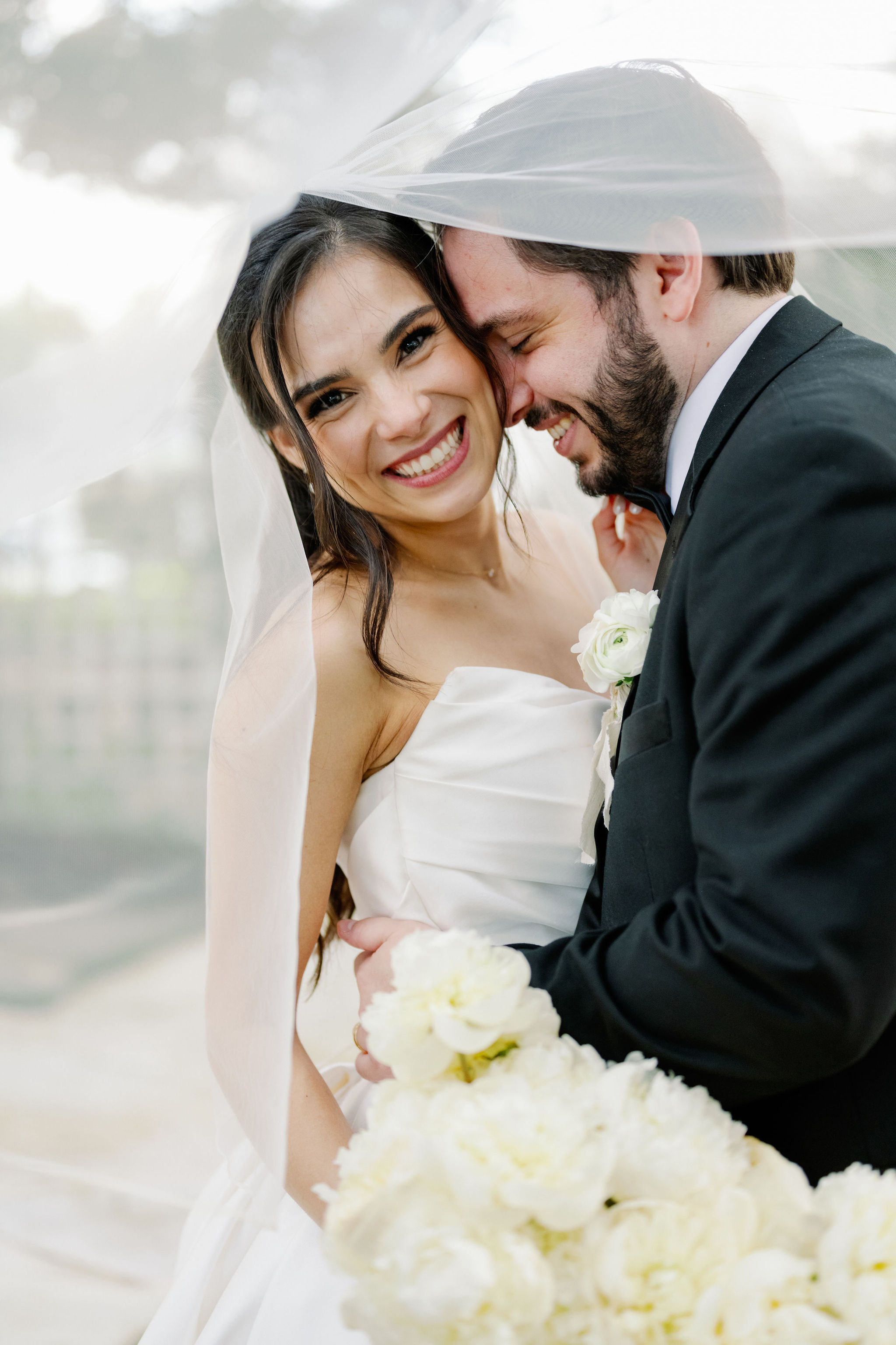 A smiling bride and groom embrace beneath a sheer veil, holding a bouquet of white peonies and wearing a white floral boutonniere.