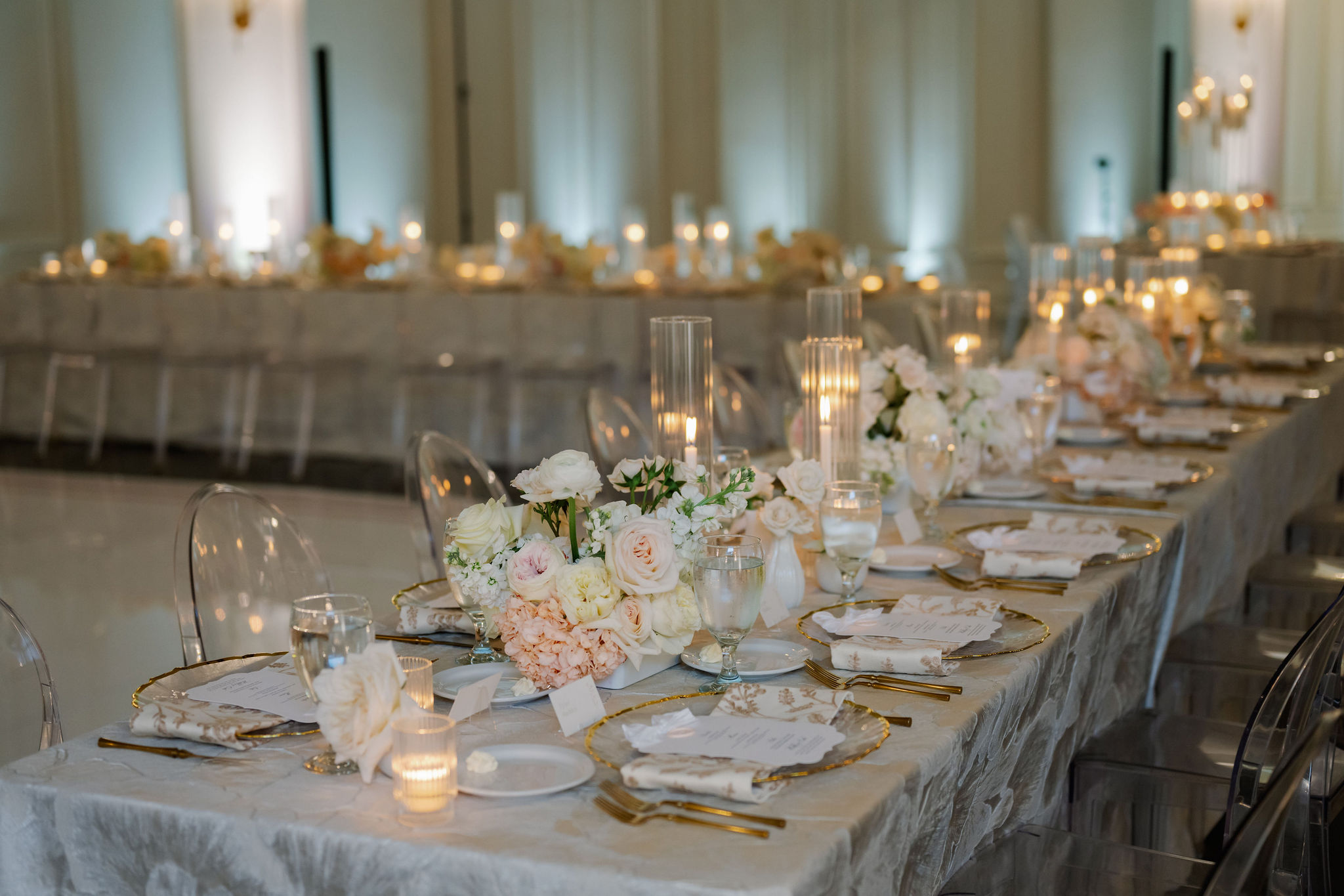 Elegant reception tablescape with blush and white floral arrangements, tall glass hurricane candles, and gold-rimmed place settings.