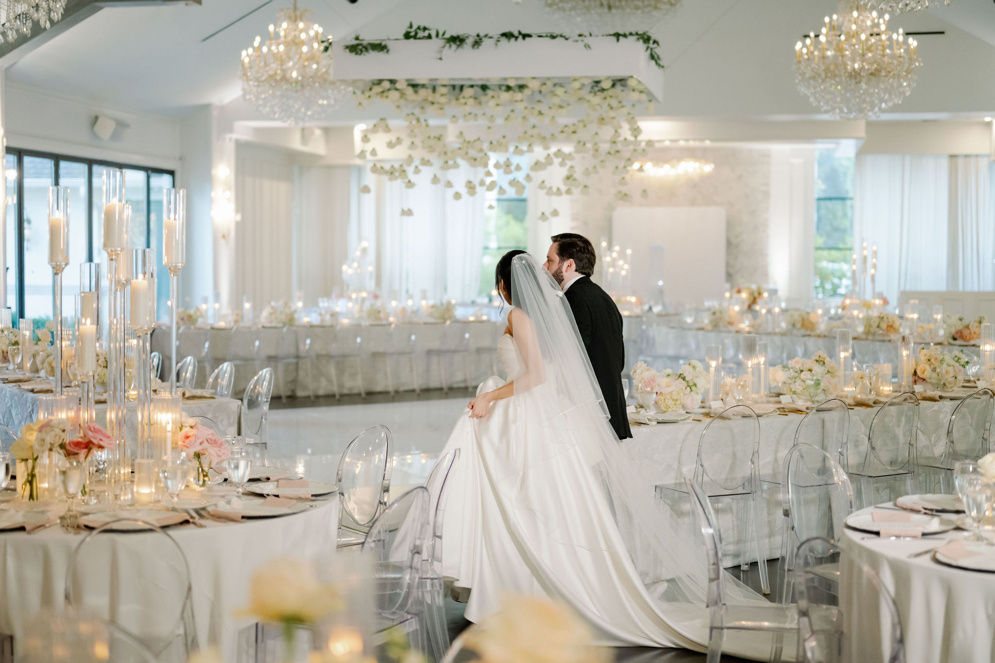 A bride and groom walk through an elegant white ballroom featuring suspended floral installations, chandeliers, and candlelit tables.