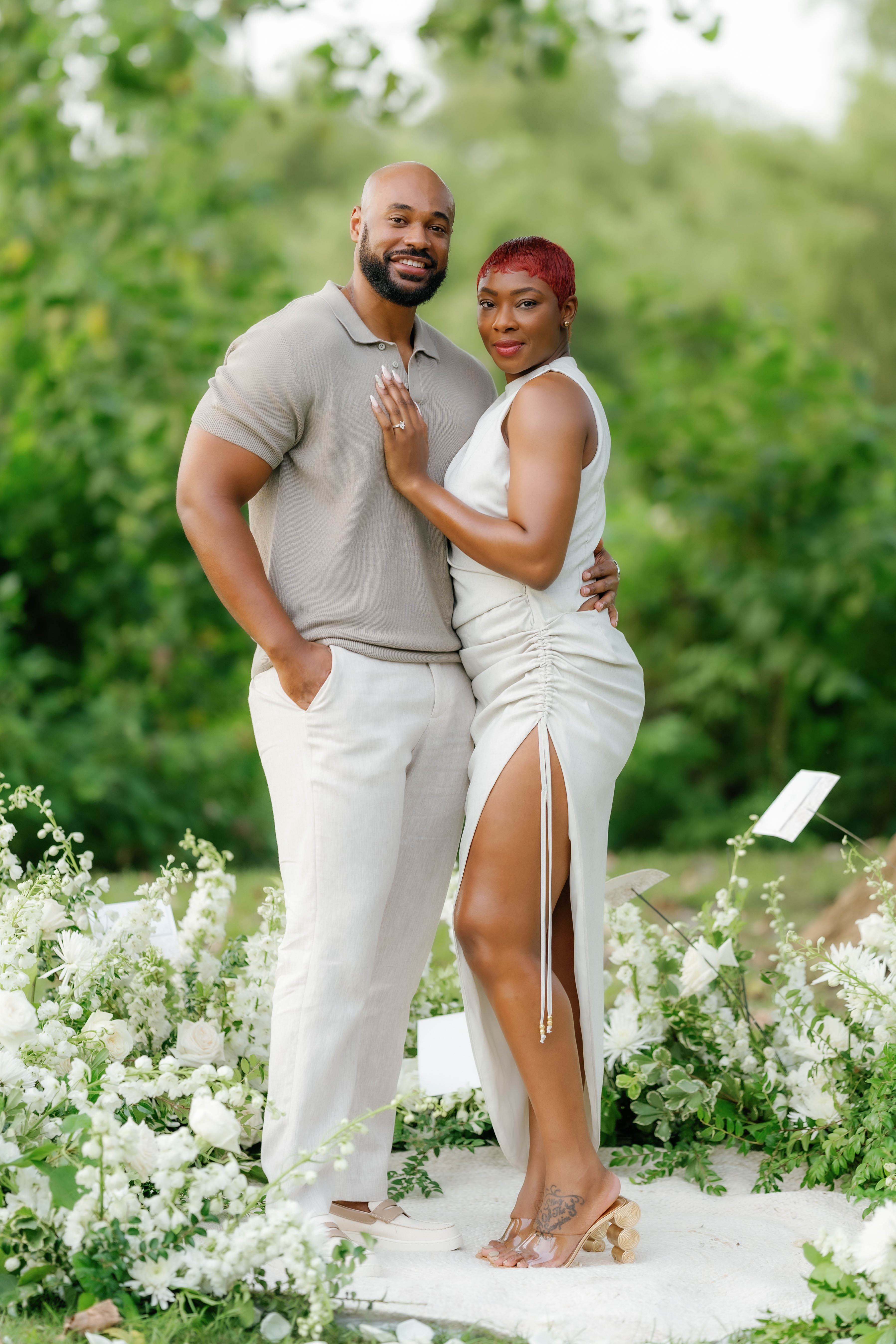 A smiling couple dressed in neutral tones embraces outdoors against a lush green backdrop.