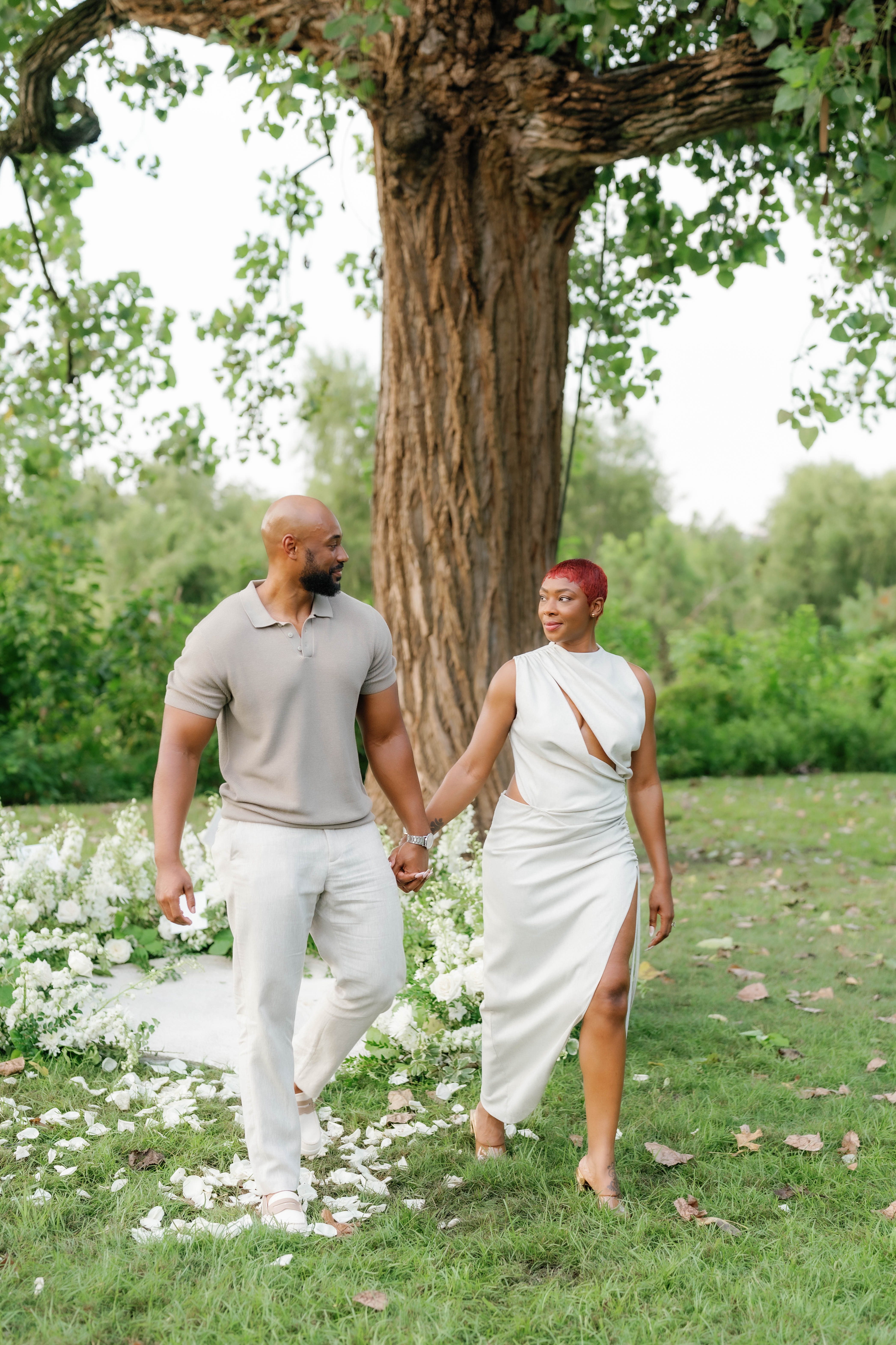 A couple holds hands outdoors on a grassy lawn scattered with white rose petals in front of a large tree.