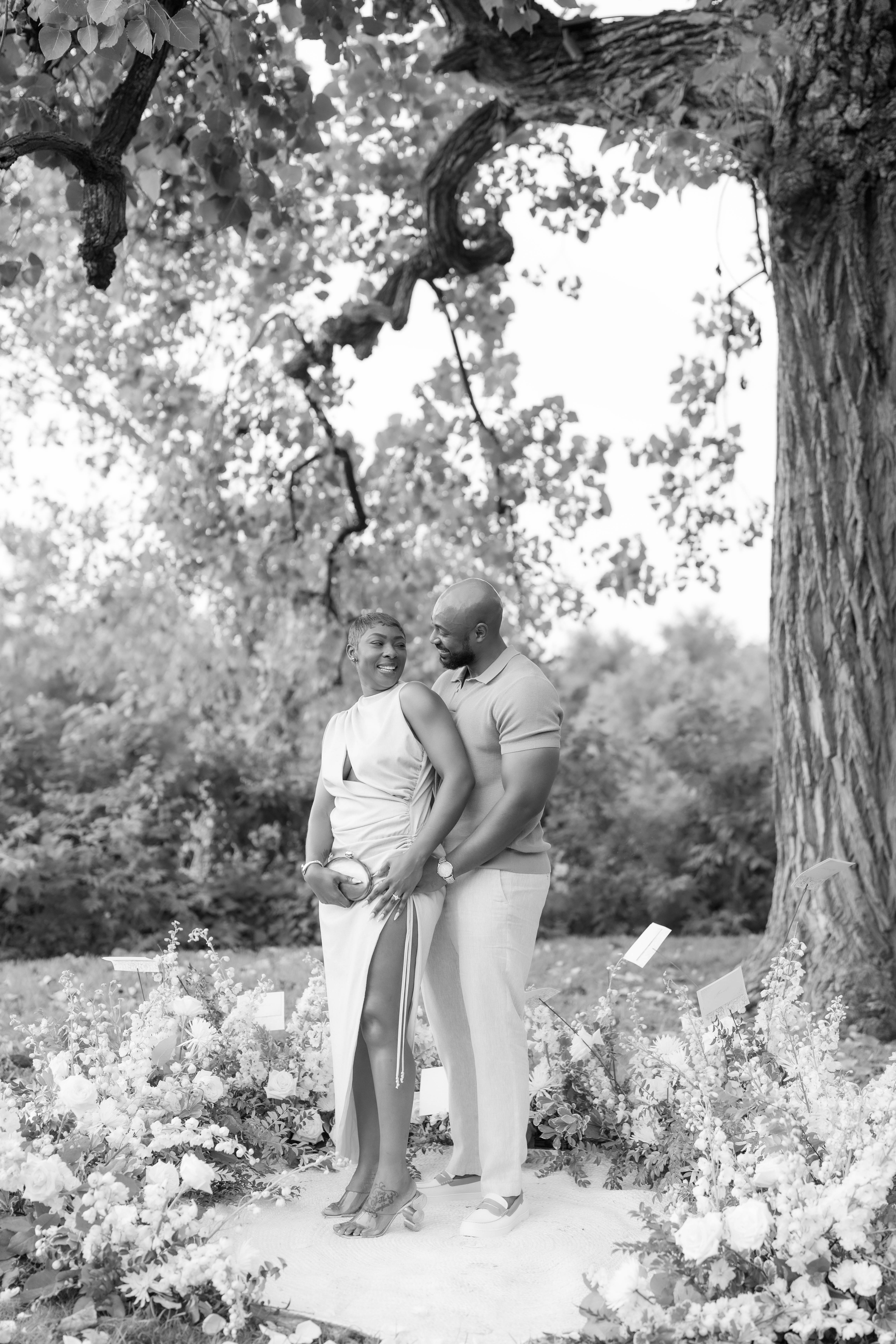 A smiling couple embraces beneath lush tree branches during an outdoor private celebration.