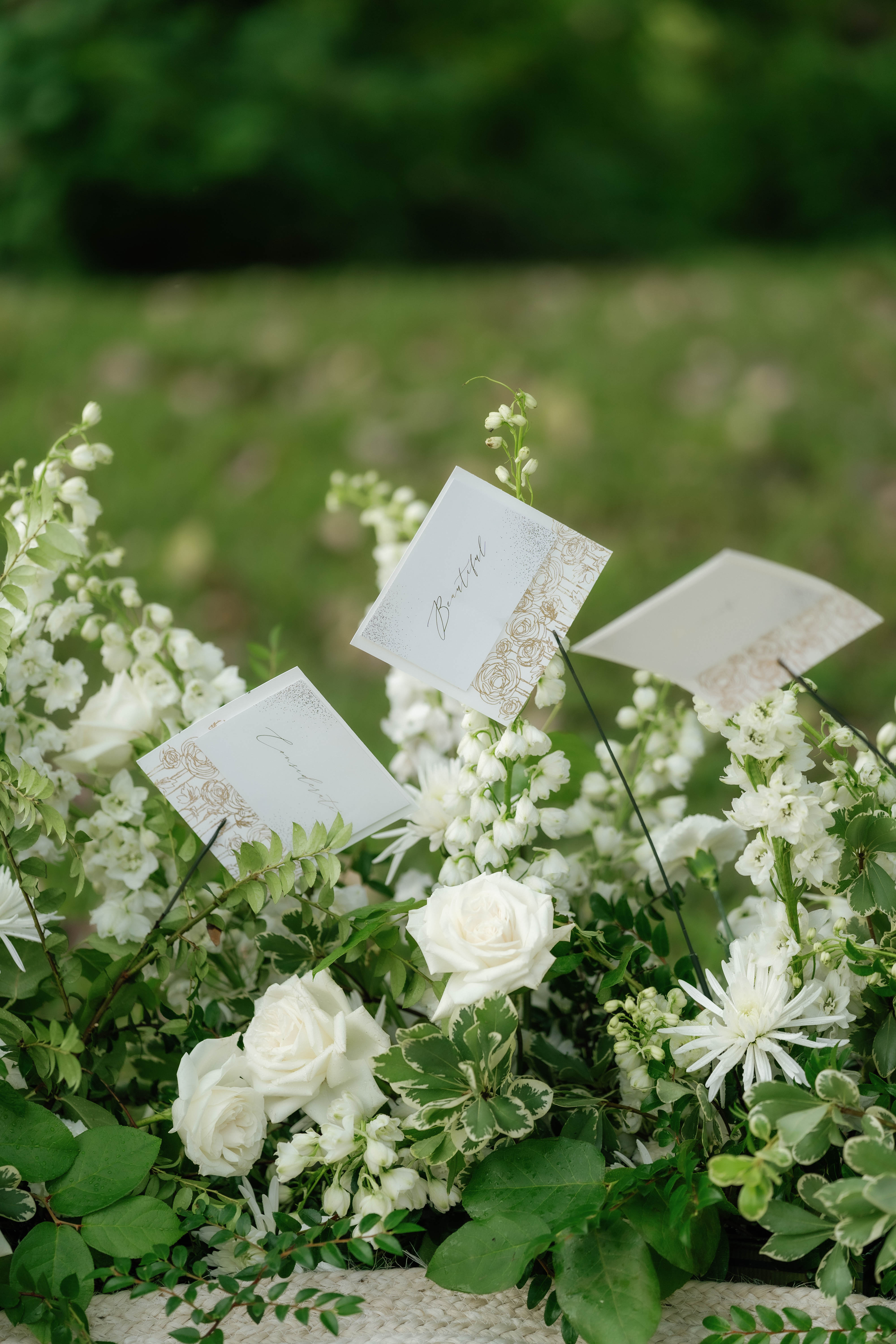 White roses, delicate bell-shaped blossoms, and lush greenery tucked beside an elegant stationery card outdoors.