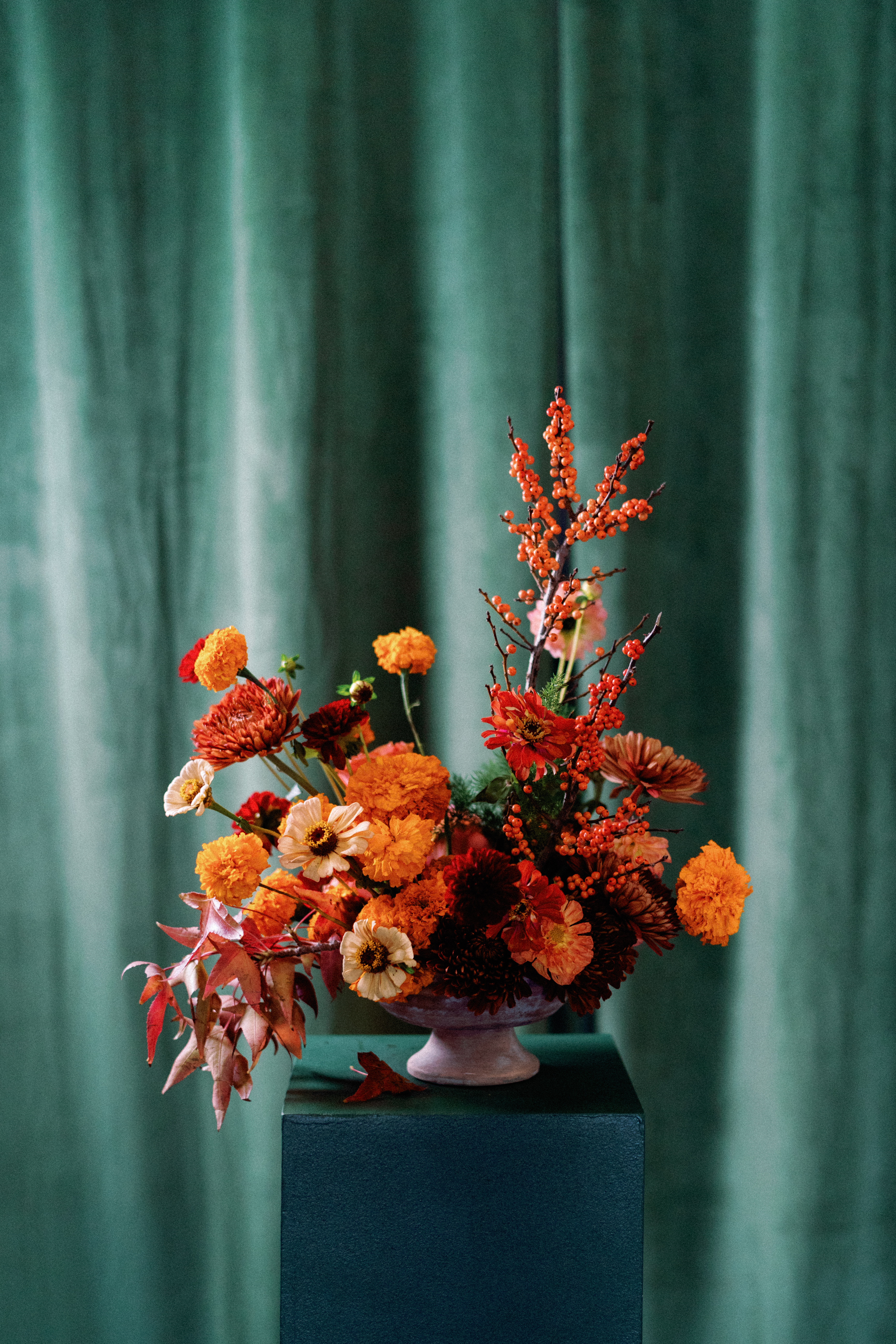 An autumn floral arrangement of orange marigolds, zinnias, and berry branches in a pedestal urn against deep green velvet drapery.