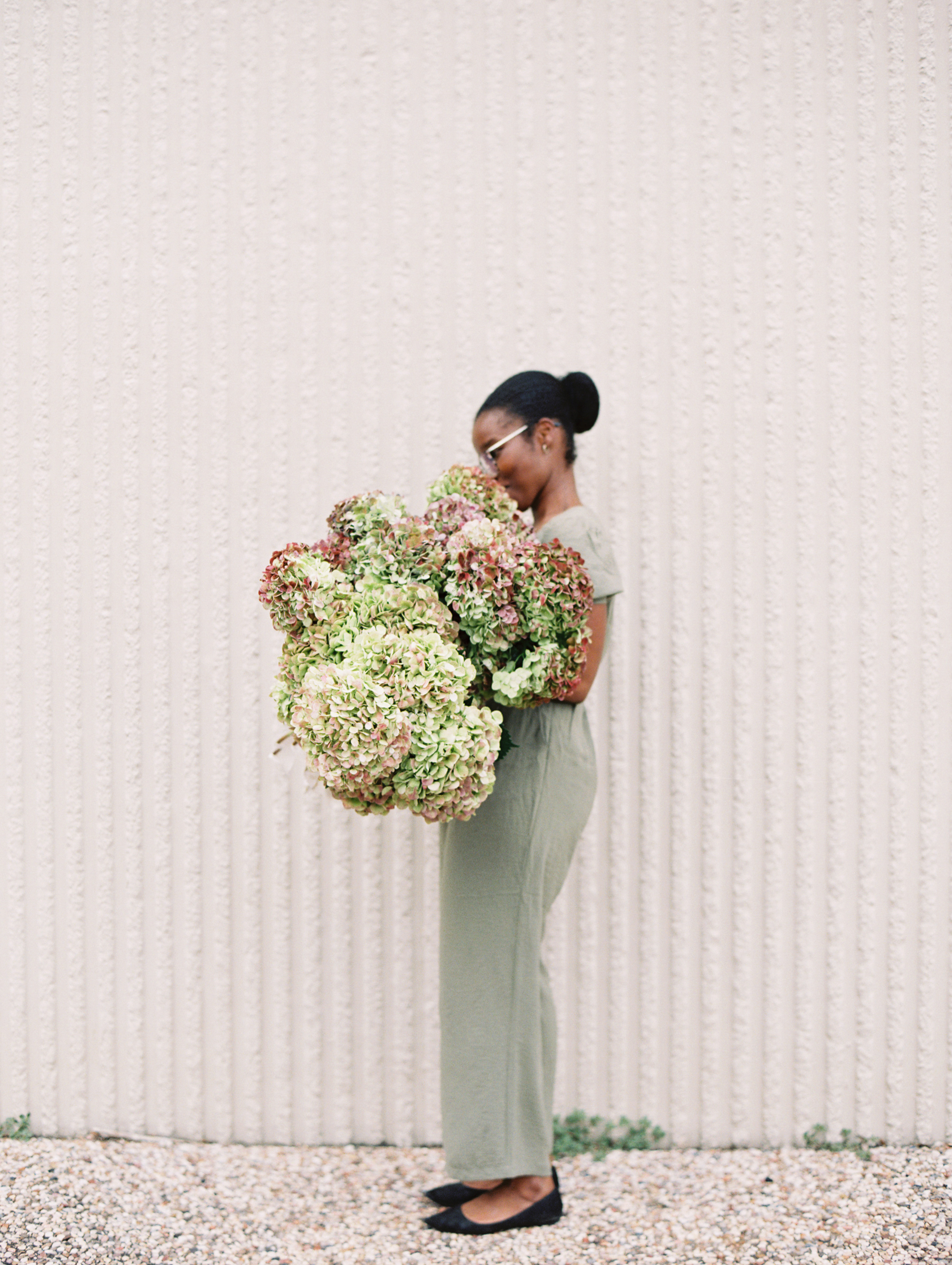 A woman in a sage green jumpsuit carries a lush armful of antique green and mauve hydrangeas.
