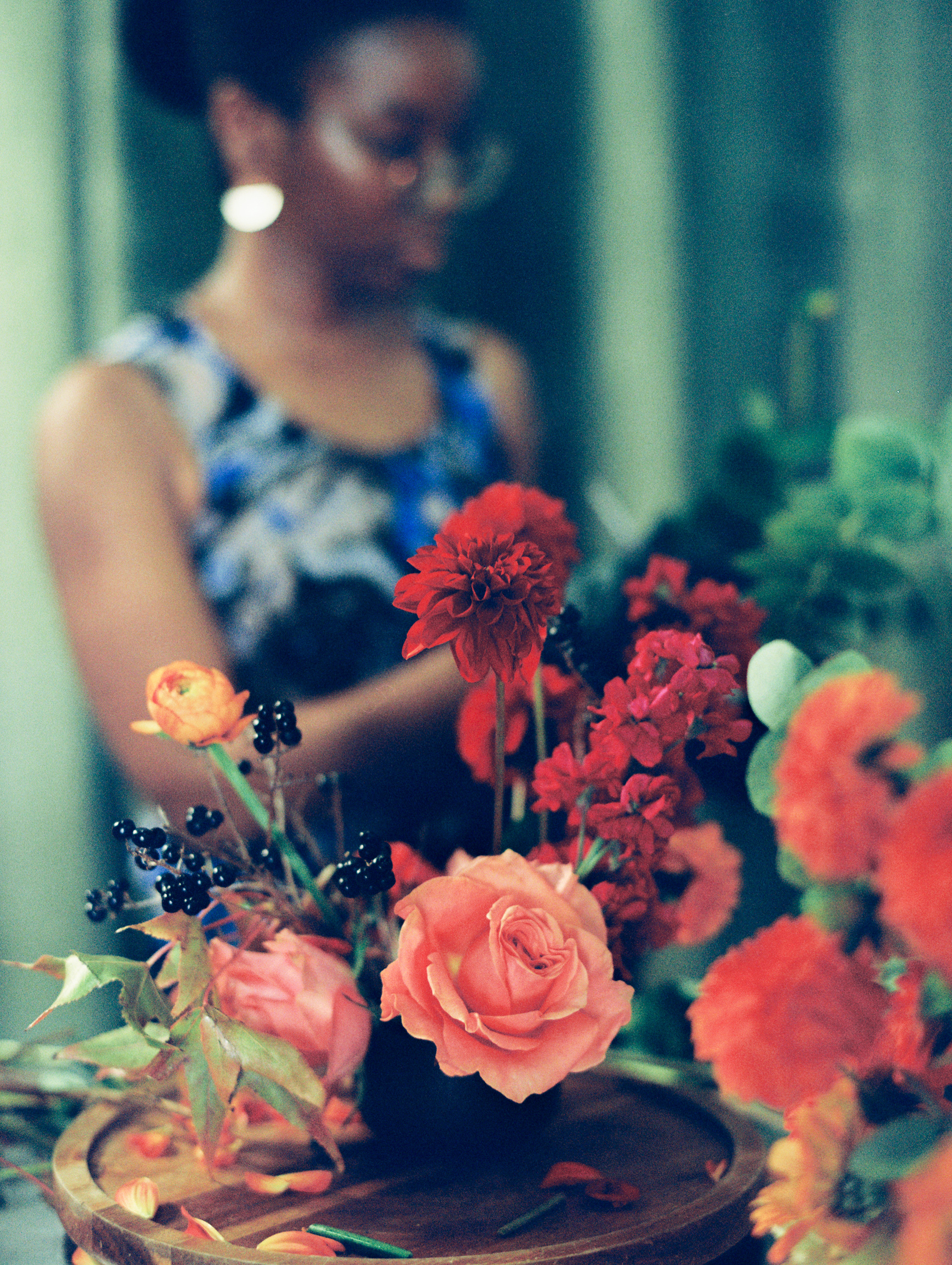 A florist arranges vibrant red dahlias, peach garden roses, and dark berries into a bespoke floral centerpiece.