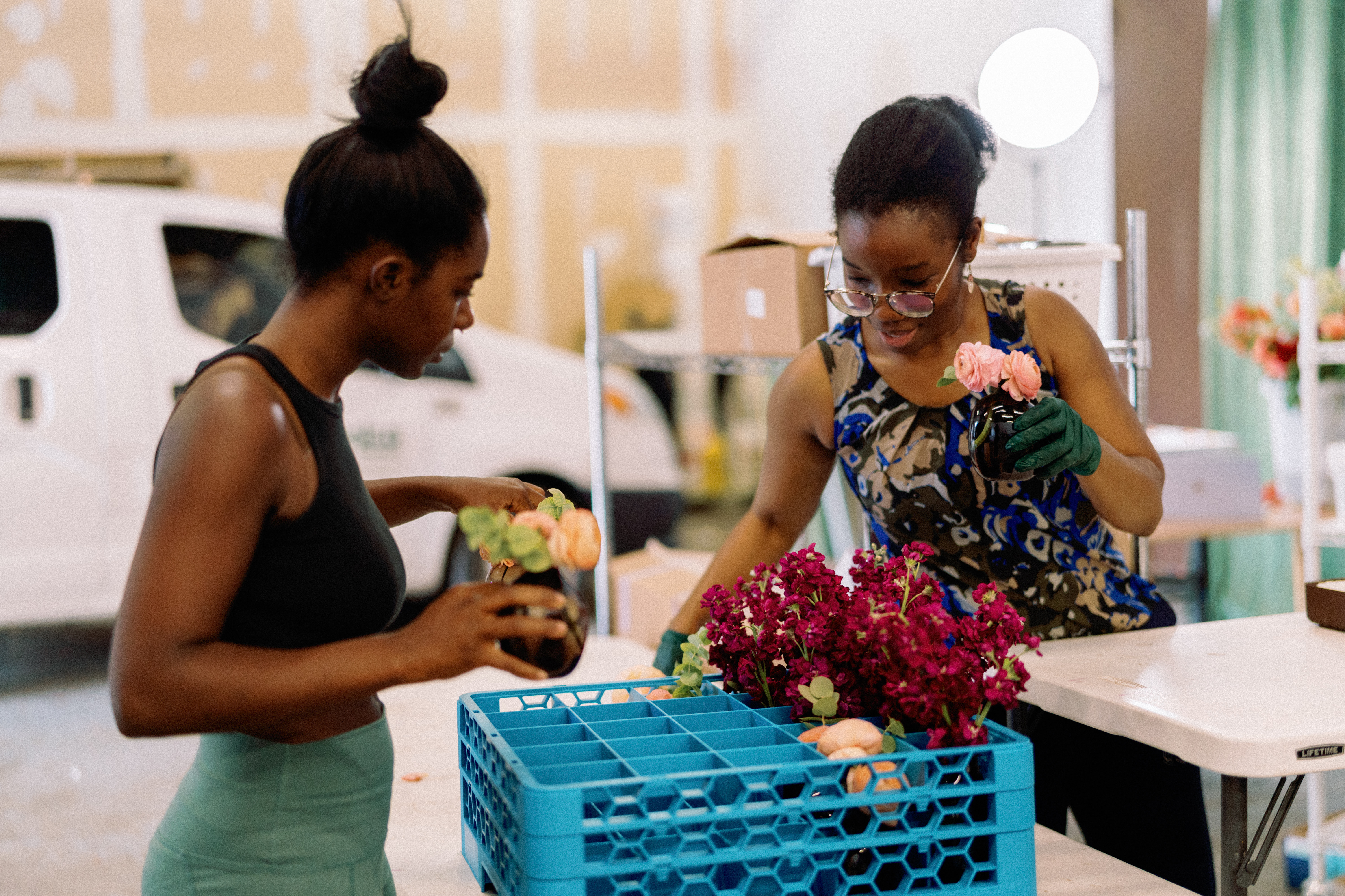 A florist prepares vibrant magenta blossoms and peach flowers in a workshop alongside a blue transport crate.