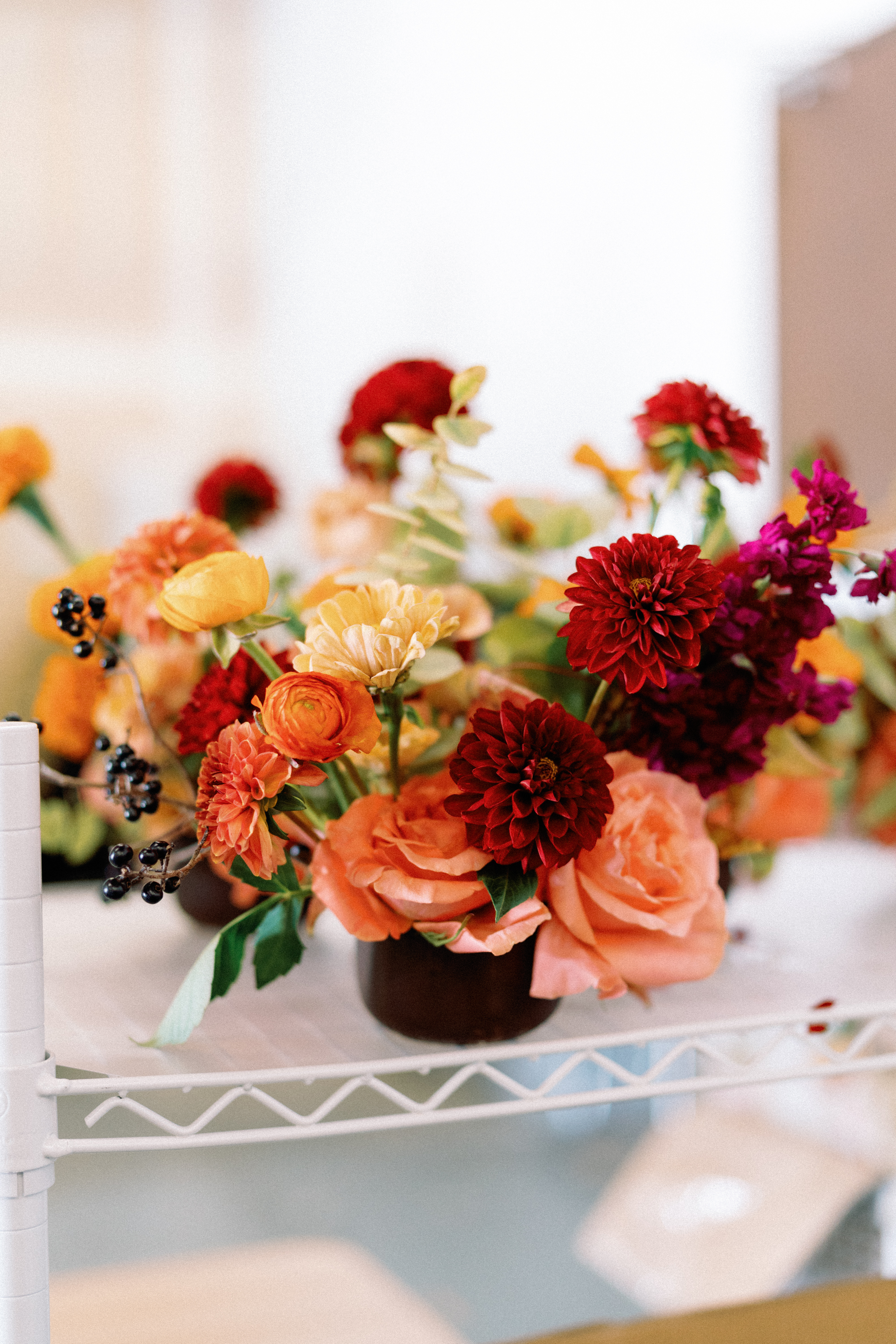 A floral arrangement of deep crimson dahlias, peach roses, and eucalyptus in a dark ceramic bowl on a studio wire shelf.