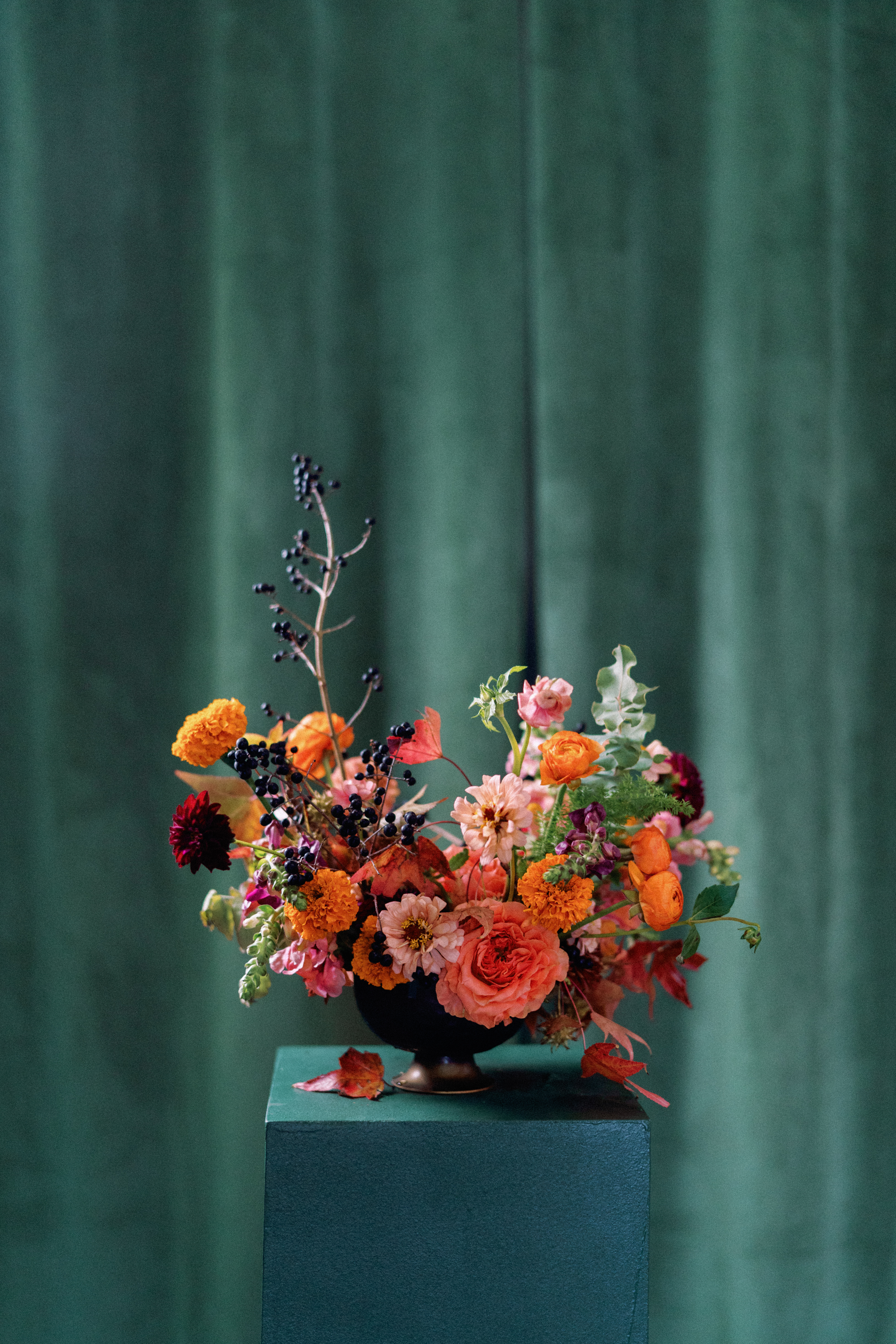 Autumn floral centerpiece with coral roses, blush blooms, and dark berries set against a rich emerald green backdrop.