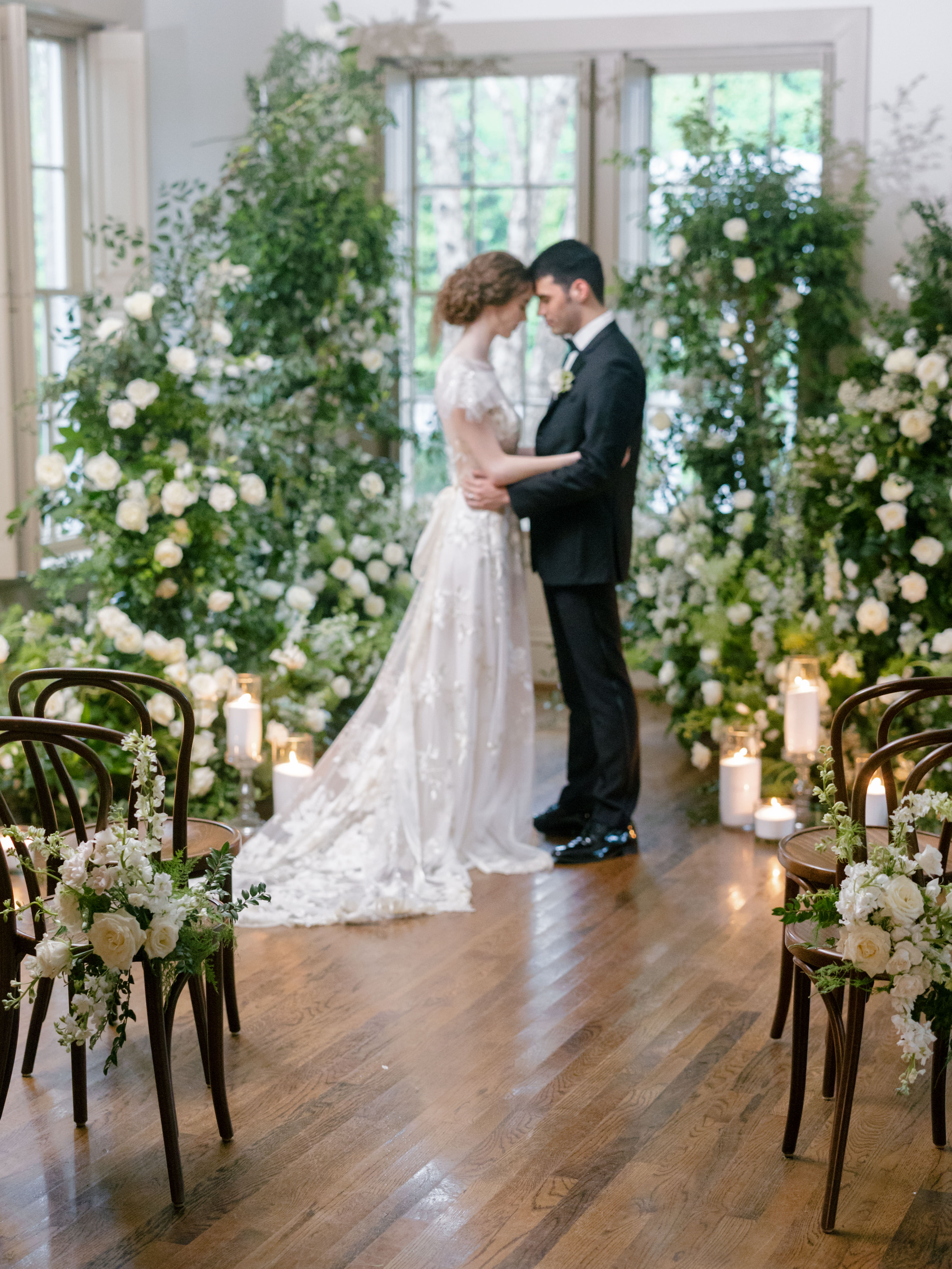 Bride and groom embrace indoors beside lush white rose and greenery floral installations by tall windows.