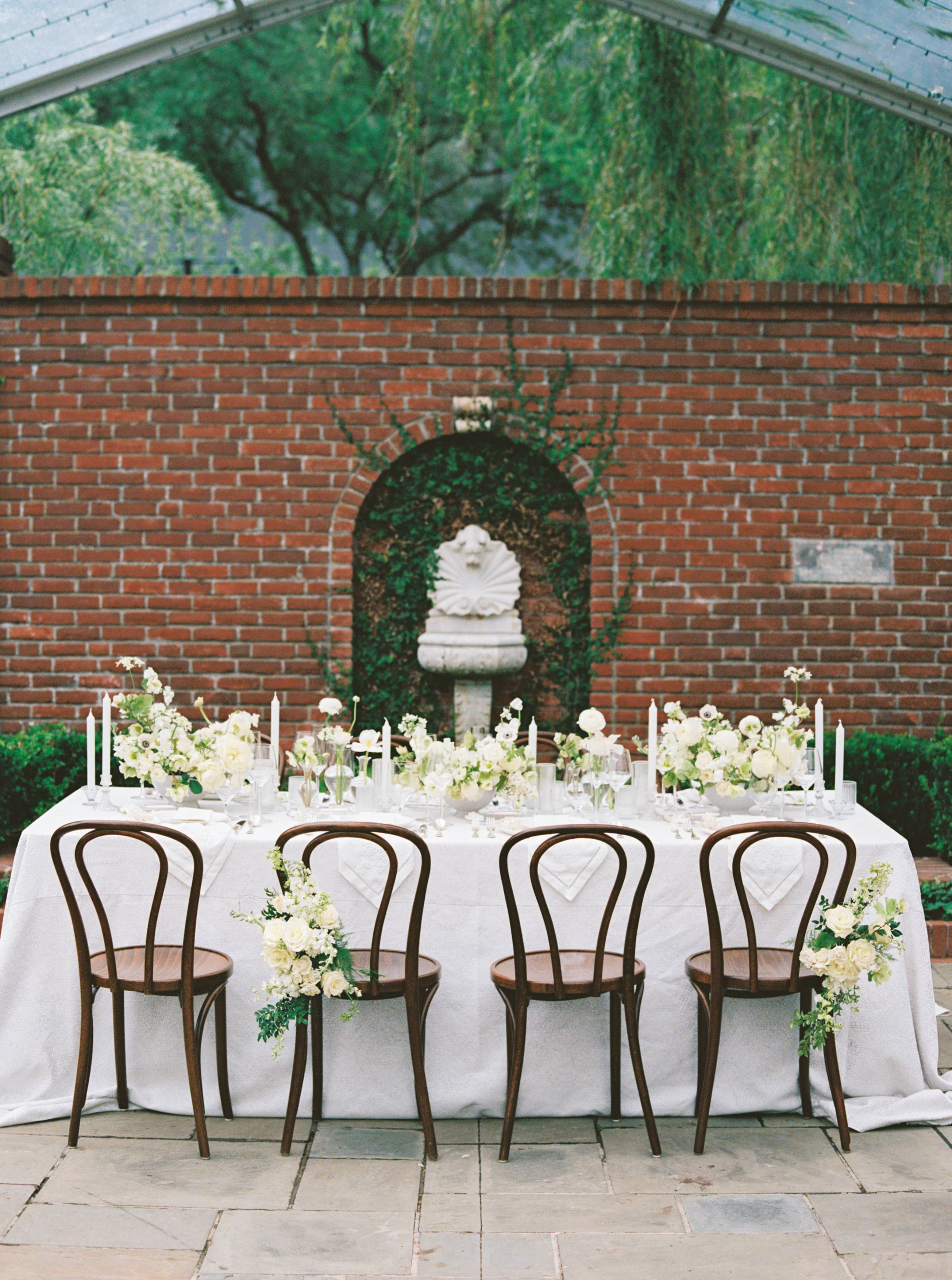 An outdoor dining table arranged with white florals, taper candles, and wooden bentwood chairs before a brick wall fountain.