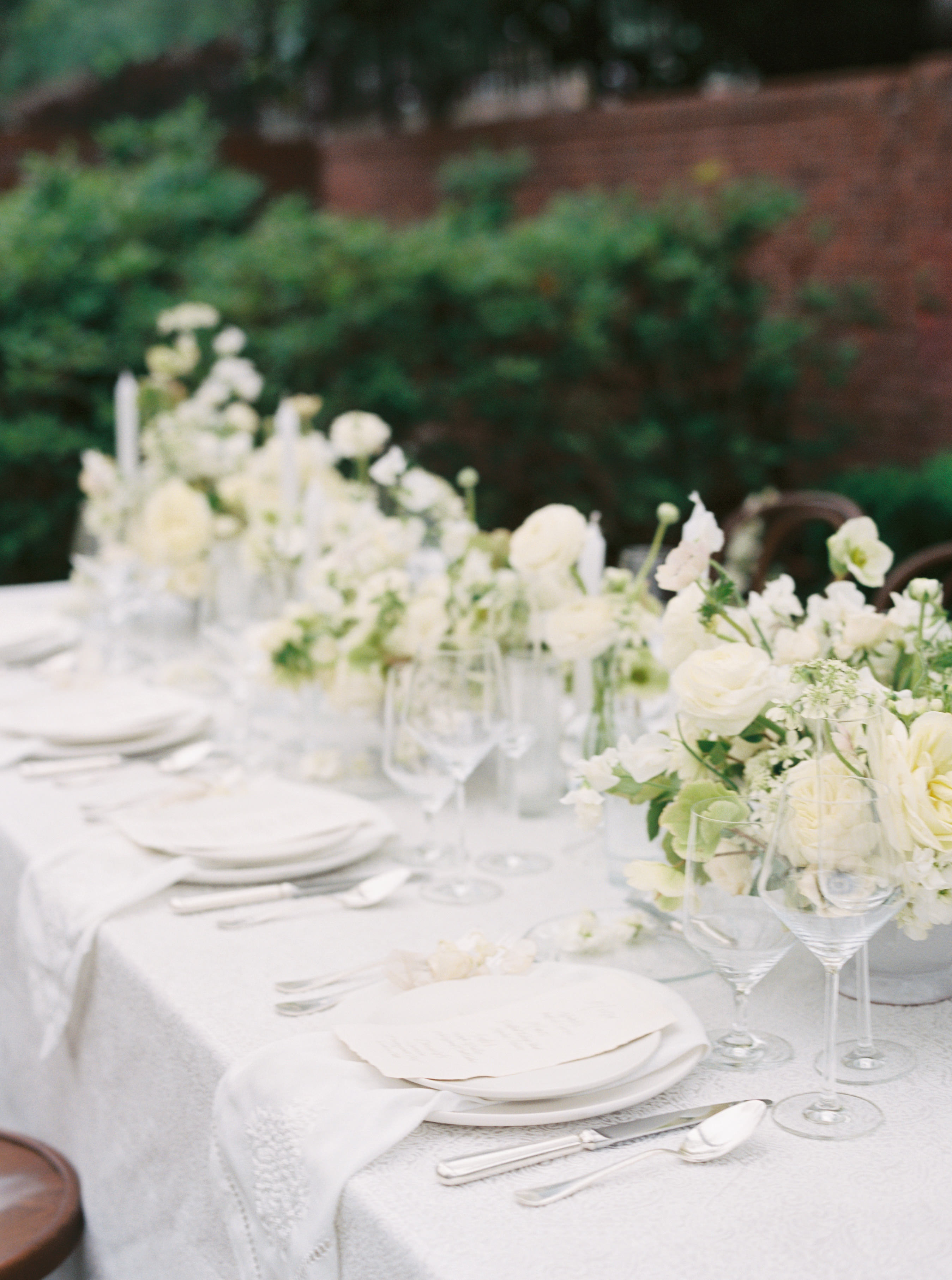 An outdoor tablescape featuring white ranunculus arrangements, fine glassware, and elegant place settings.