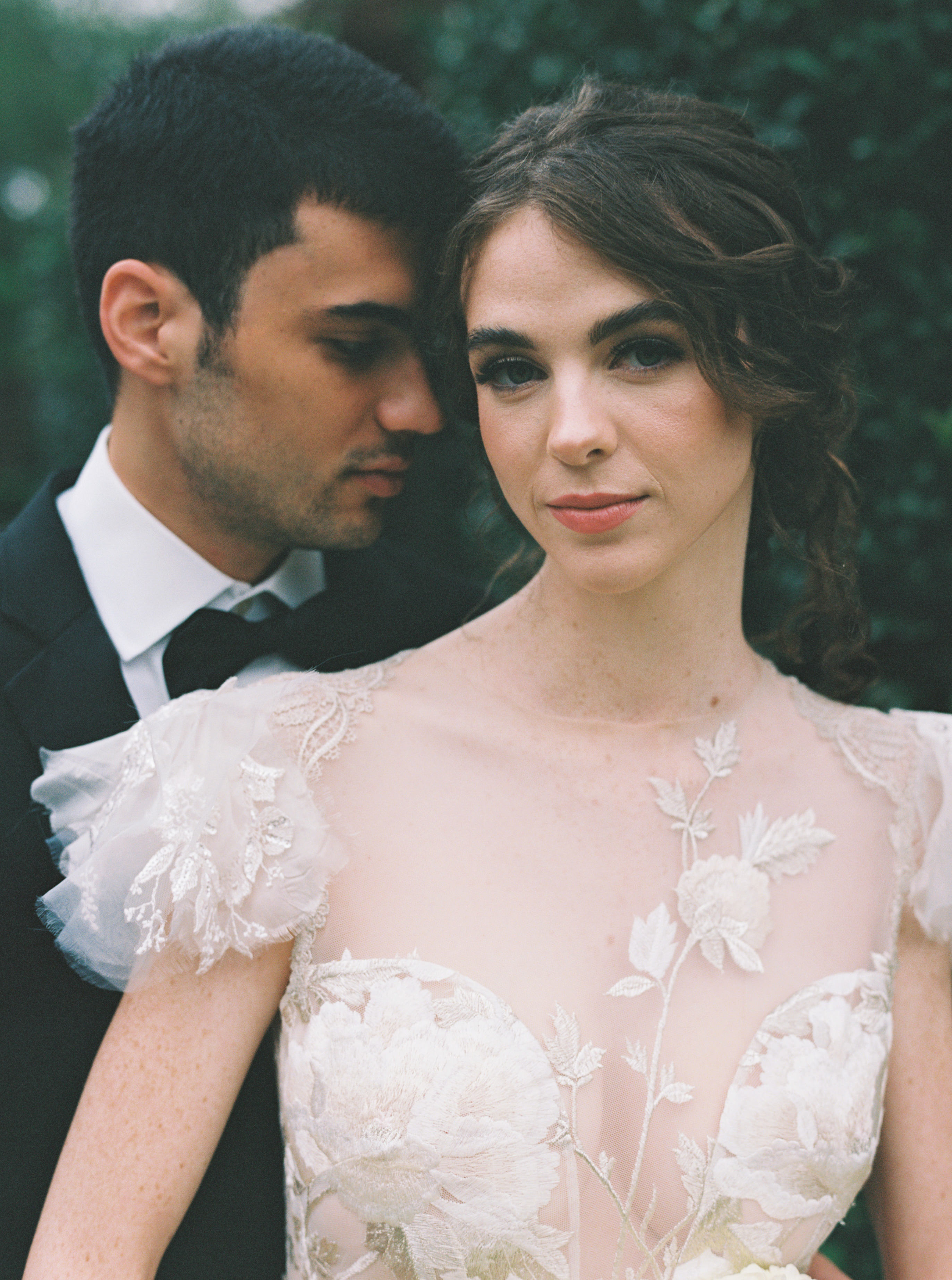 A bride in an embroidered floral lace gown poses intimately with a groom in a dark suit.
