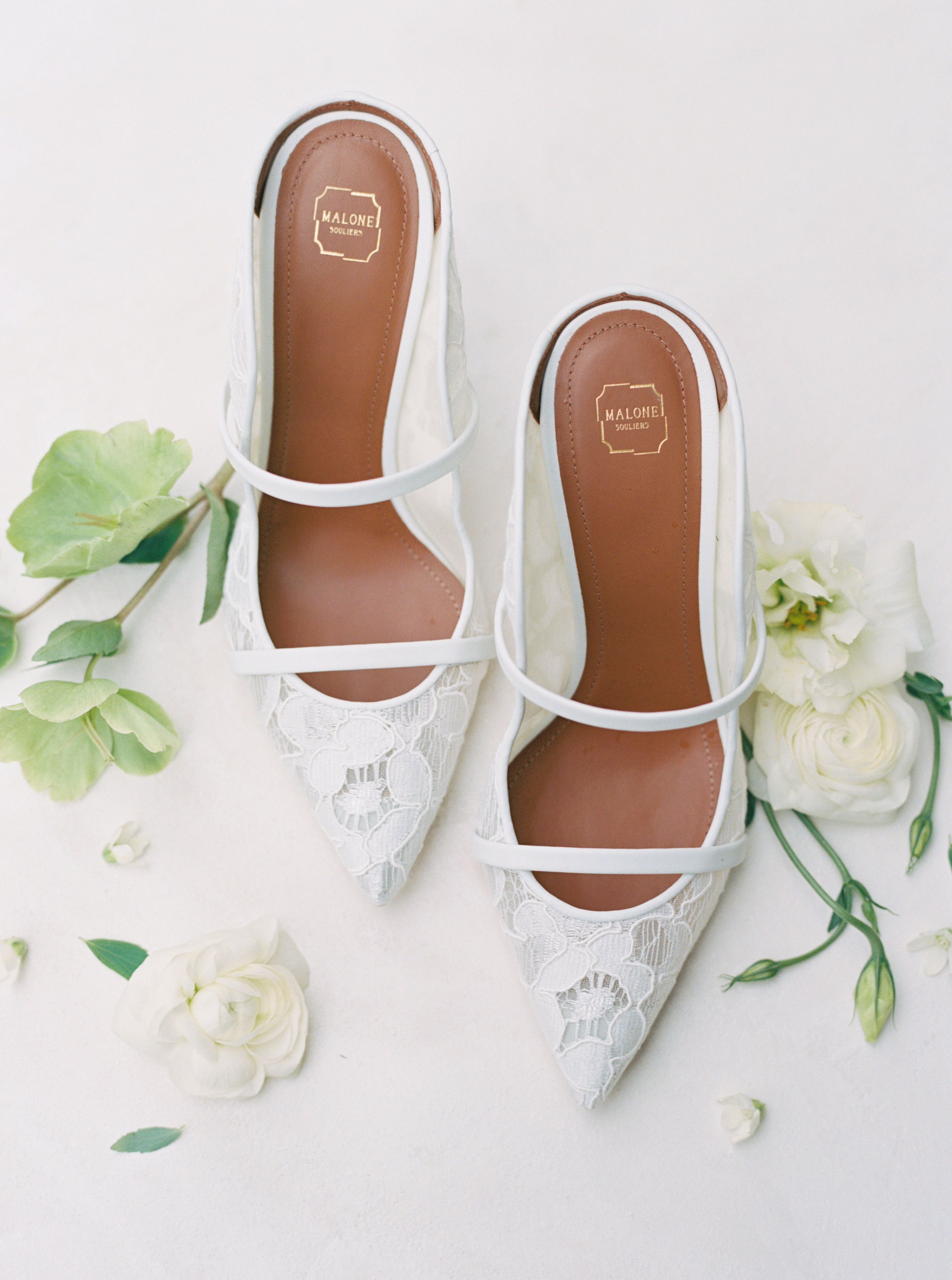 White lace pointed-toe mules with delicate straps rest on a white surface beside a small ivory blossom.