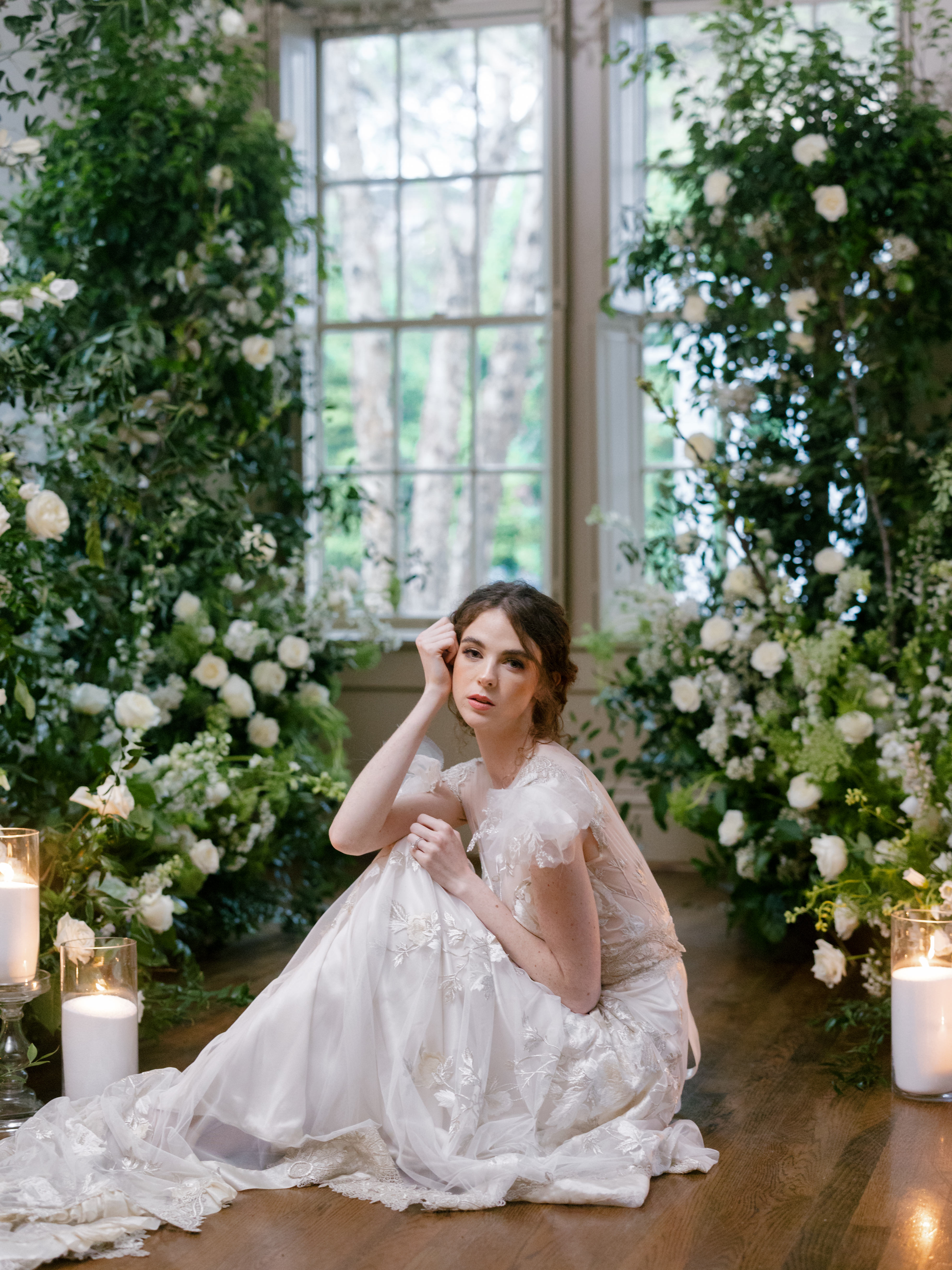 A bride in an embroidered tulle gown rests beside a tall window framed with delicate floral and greenery accents.