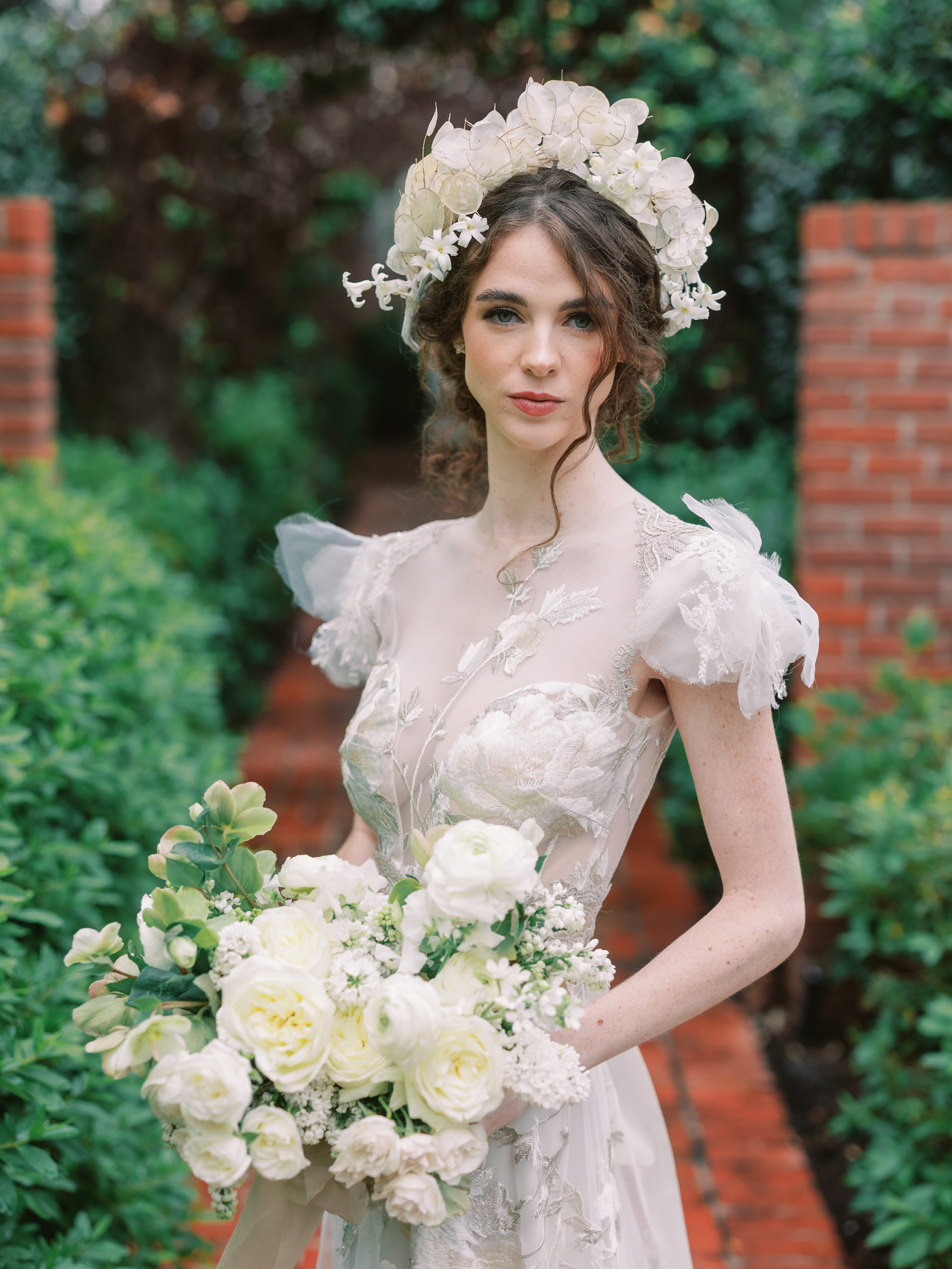 A bride in an embroidered gown and a lunaria flower crown holds a lush bouquet of white roses and lilacs.