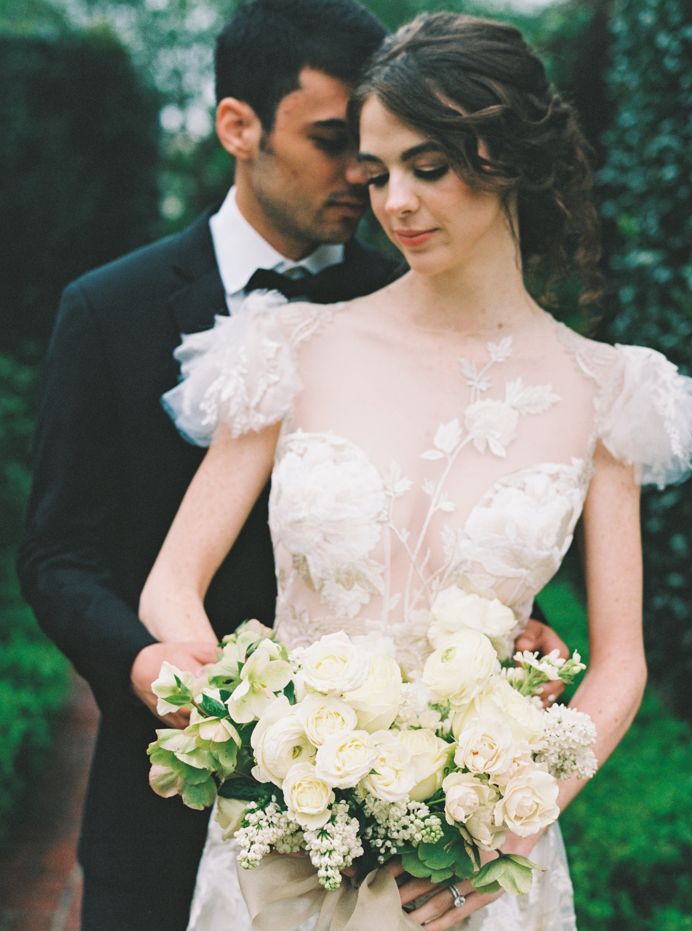 A bride in an embroidered gown holds an ivory bouquet of garden roses, ranunculus, and lilacs alongside the groom.
