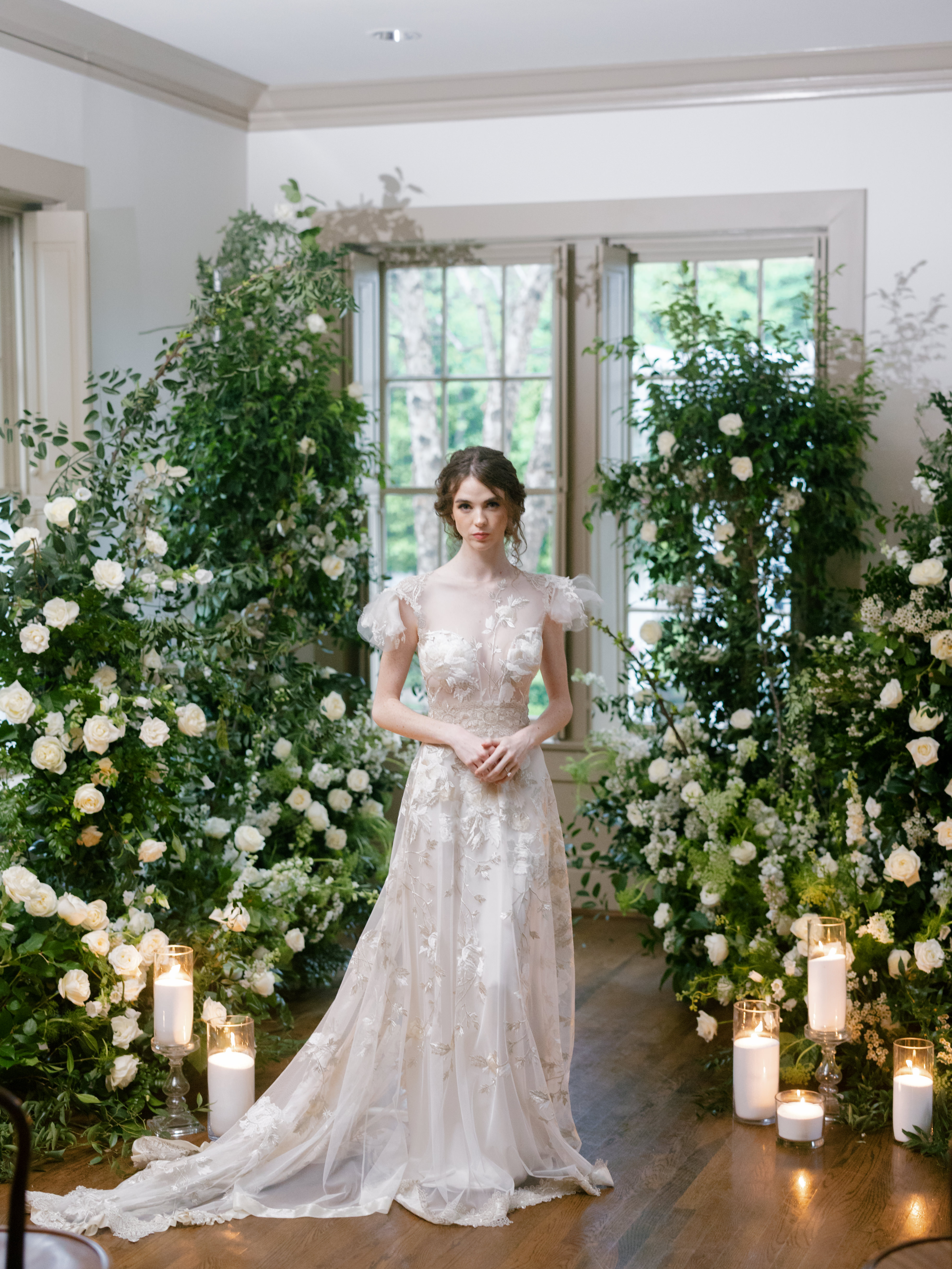 A bride in an embroidered lace gown stands between lush white floral installations before tall sunlit windows.