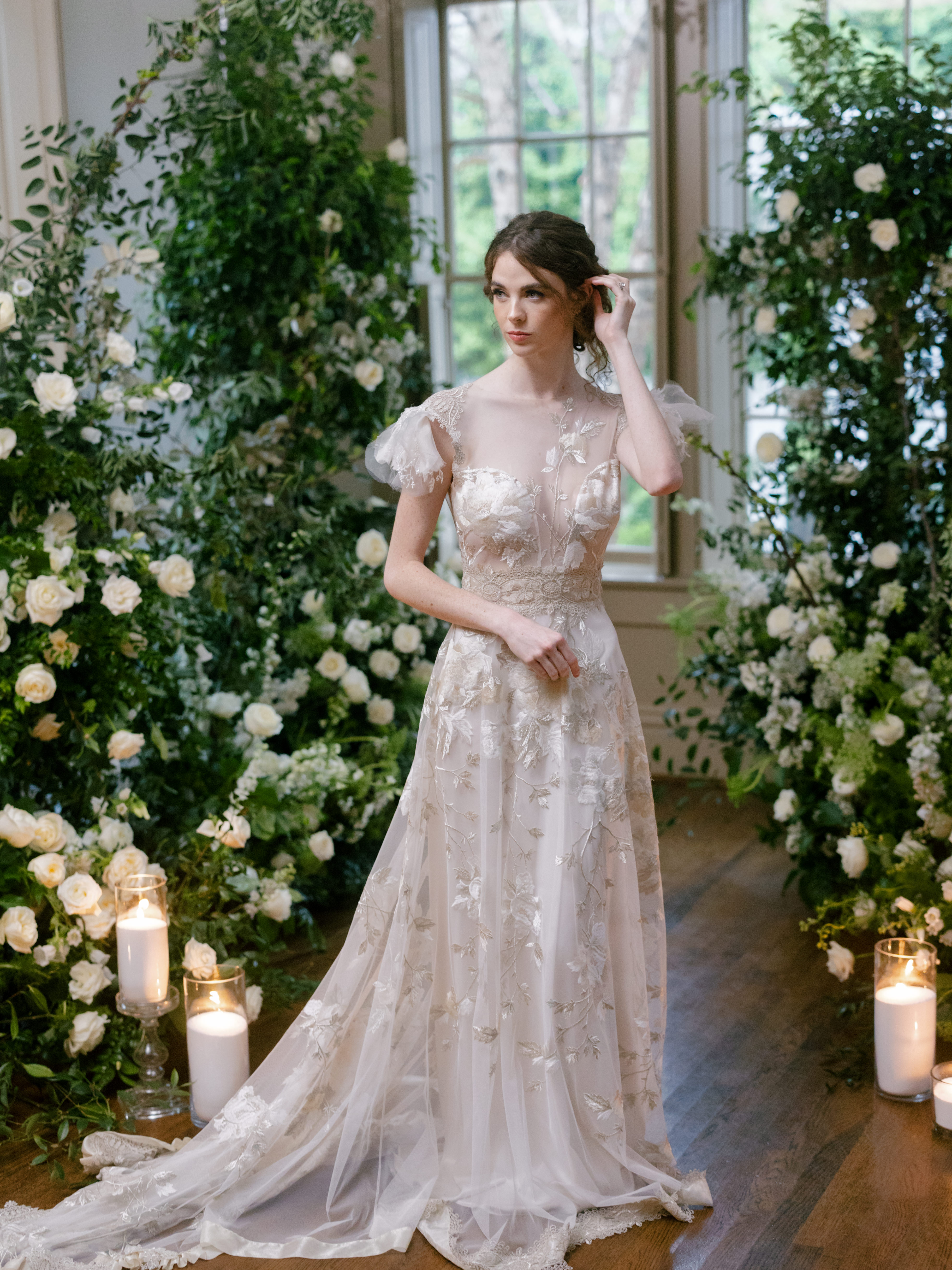 A bride in an embroidered lace gown stands beside a tall floral installation of white roses and lush greenery near large windows.