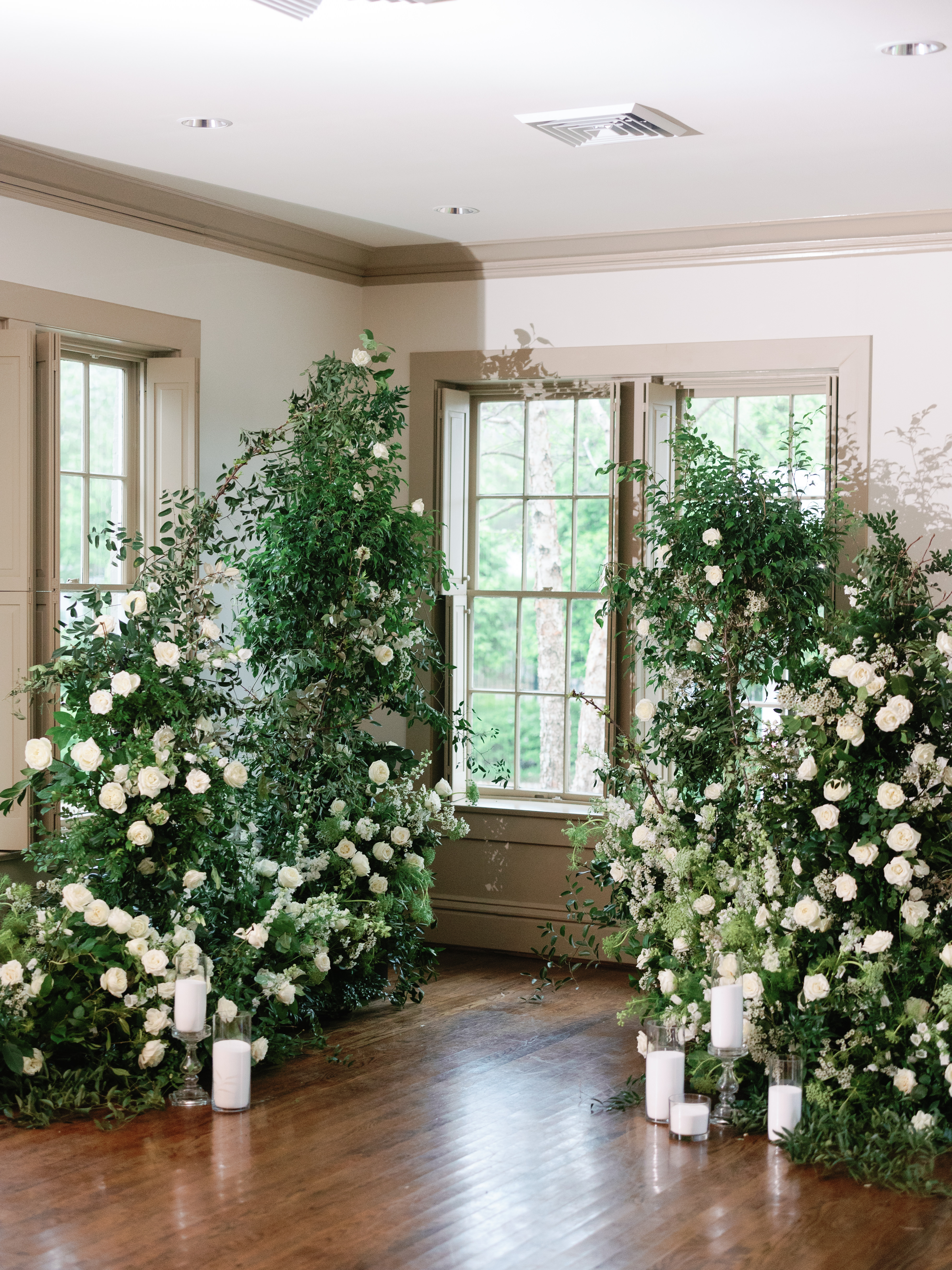 Lush greenery and white roses frame a tall indoor window overlooking trees in an elegant room.
