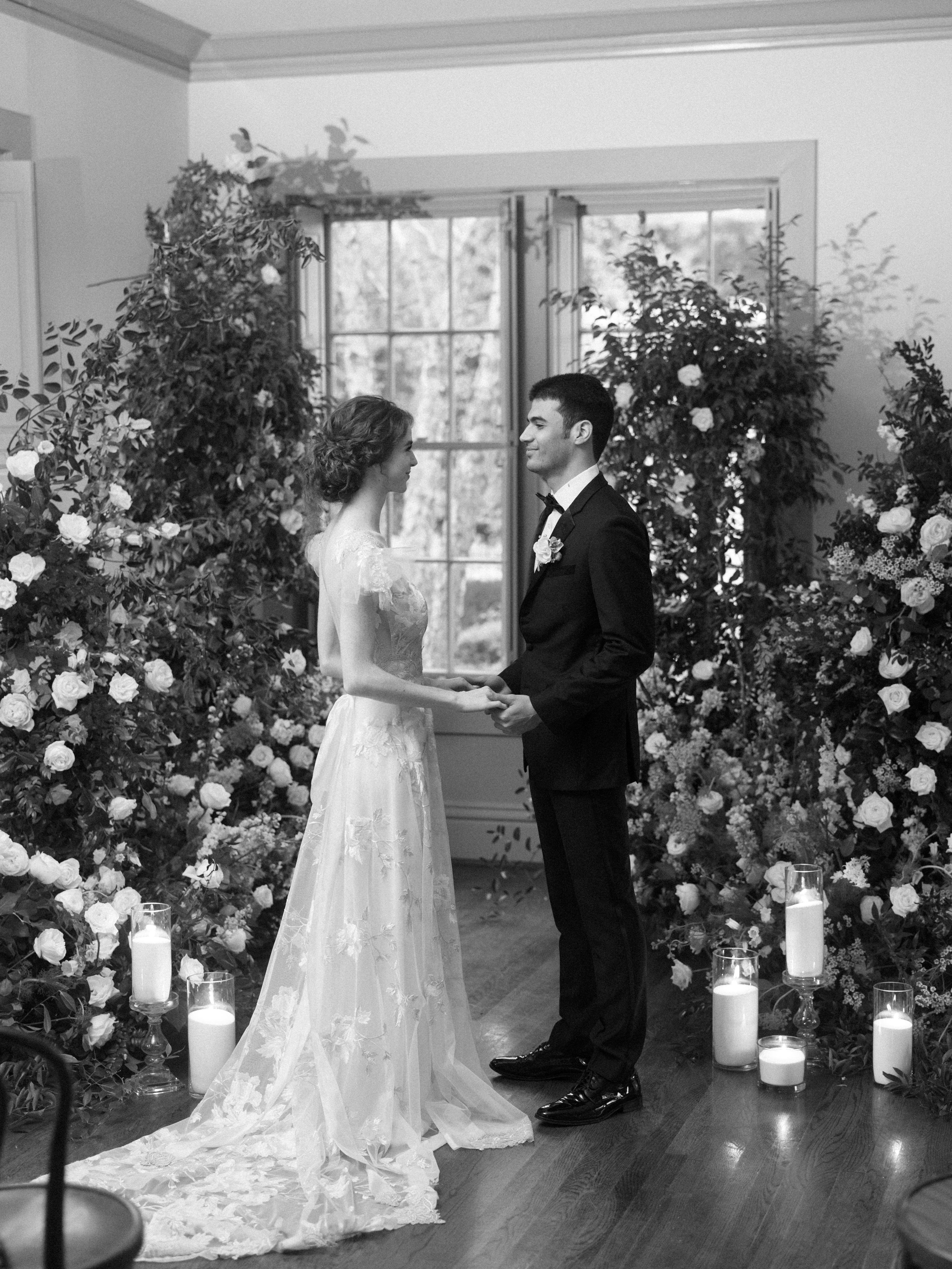 Bride and groom holding hands before tall paned windows framed by ceremony greenery in a black and white portrait.