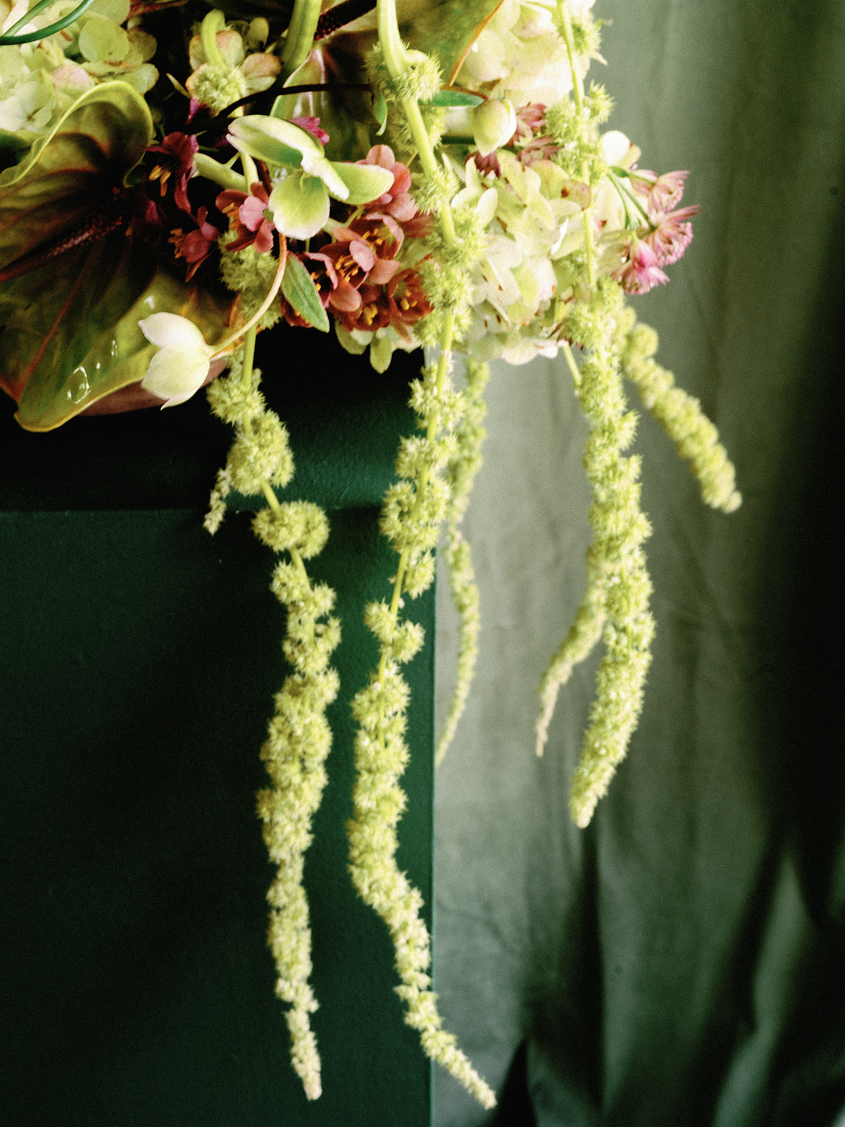 Cascading green amaranthus, hellebores, and anthuriums arranged on a dark green pedestal against draped fabric.