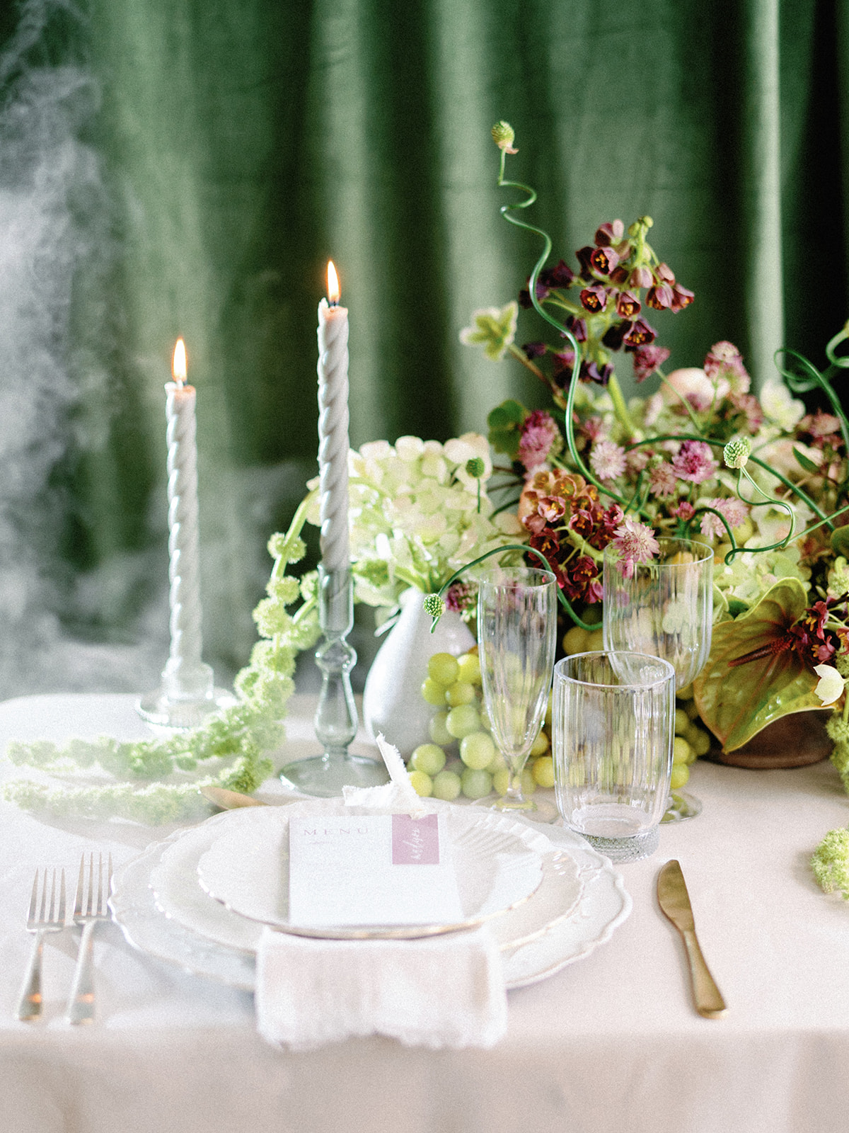 An elegant tablescape featuring twisted taper candles, scalloped tableware, and a whimsical floral centerpiece with green grapes.