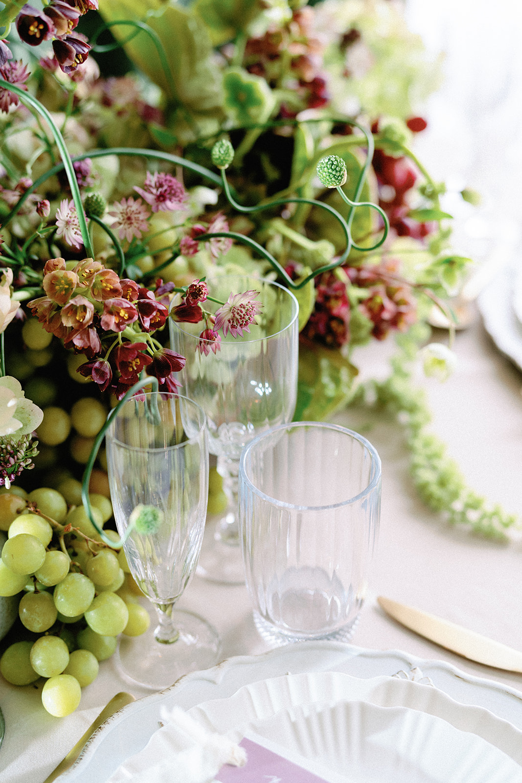 Glassware and scalloped plates set beside green grapes and a lush floral centerpiece with burgundy and mauve blooms.