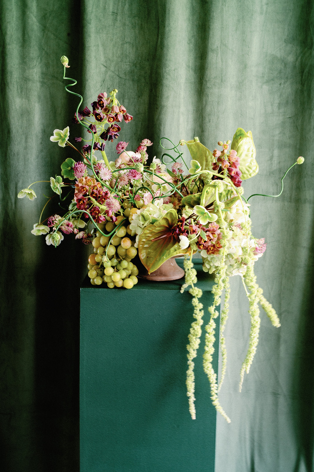 Artful floral arrangement with green grapes, hanging amaranthus, and anthuriums displayed on a green pedestal.