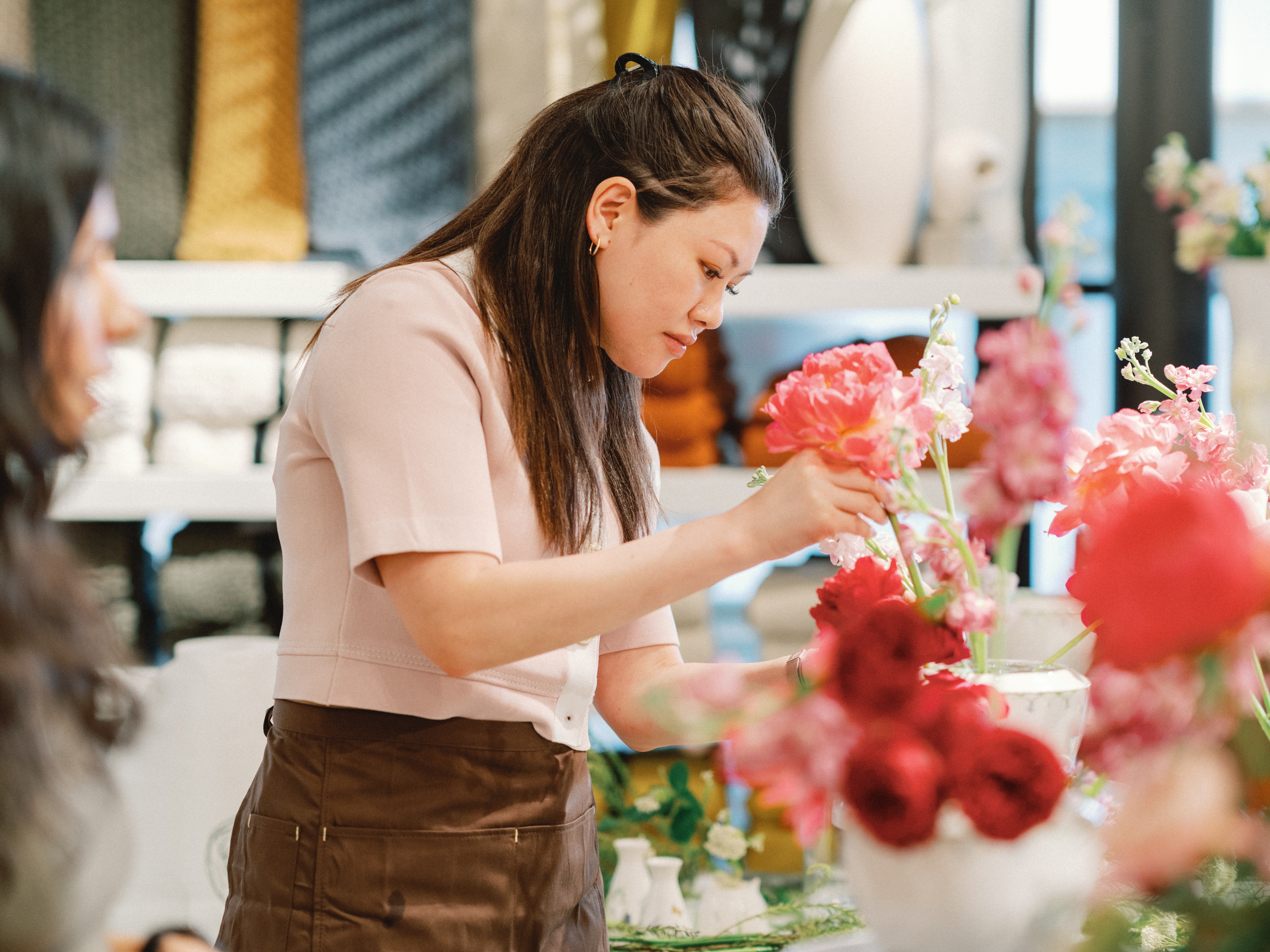 A woman wearing a brown apron attentively arranges pink flowers and greenery during an interactive floral design workshop.