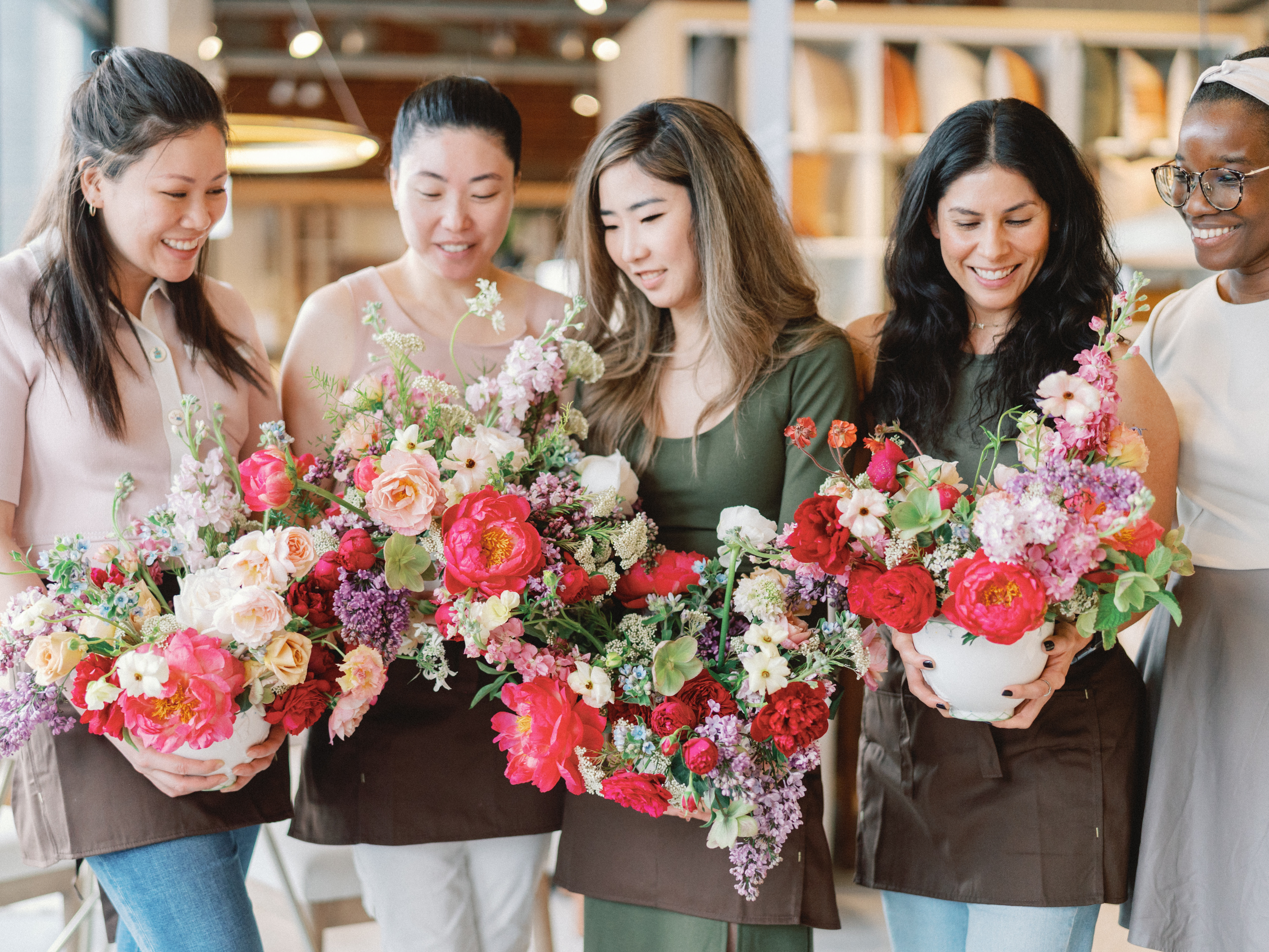 Two women smile while holding a vibrant floral arrangement filled with bright coral peonies, red roses, and lilacs.