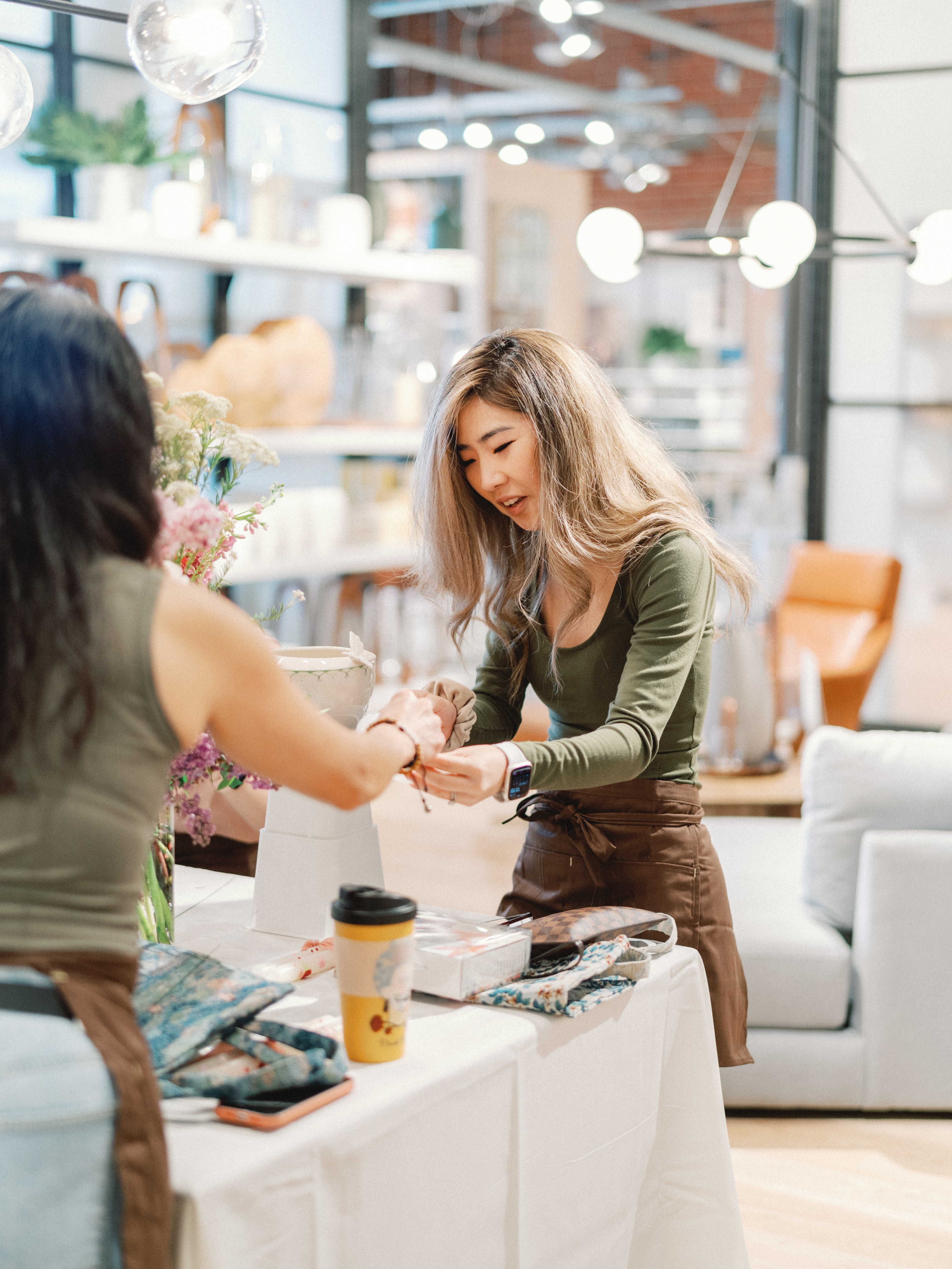 A smiling workshop host in an apron assists a guest across a table during an indoor creative session.