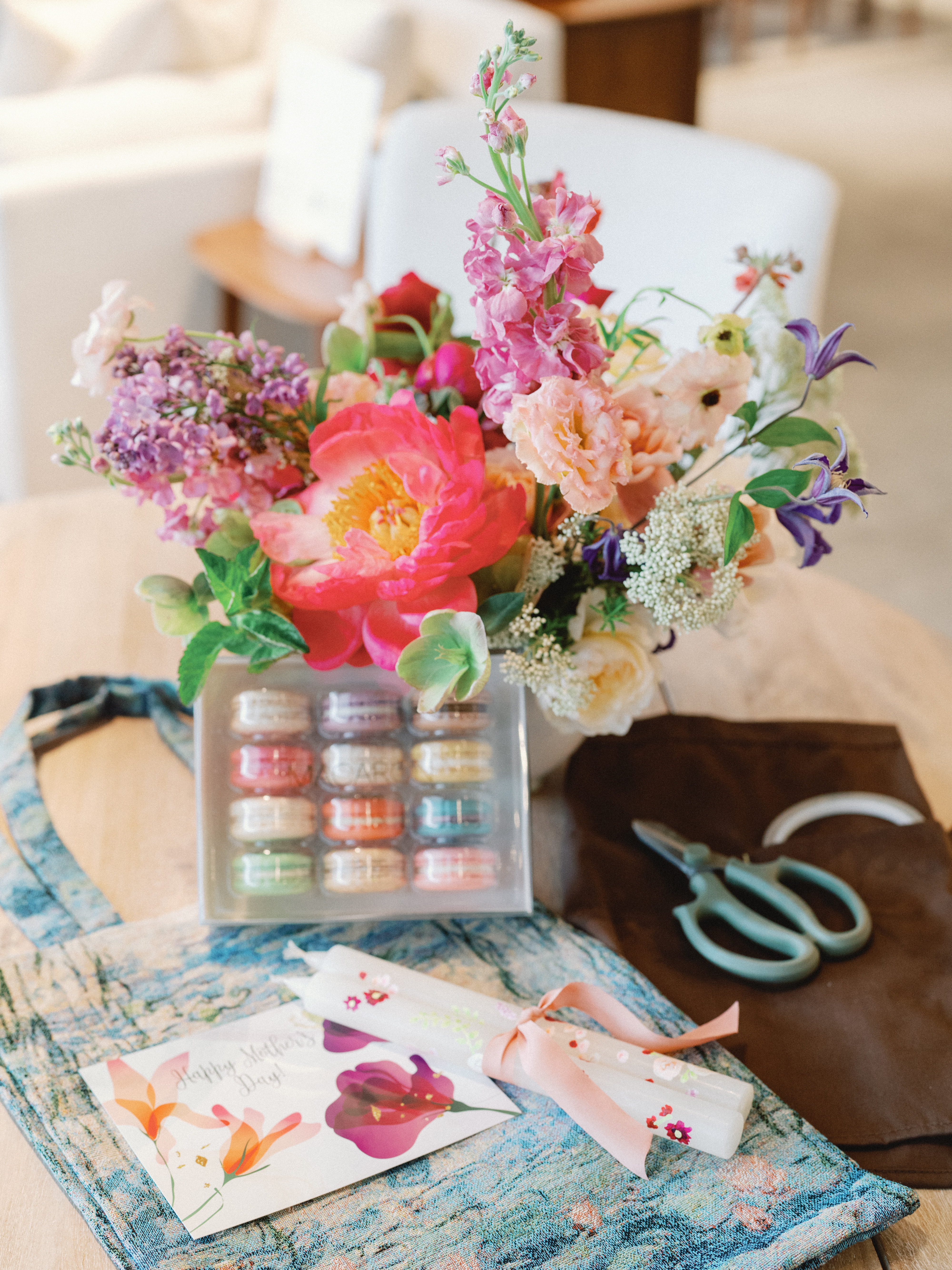 A vibrant floral arrangement with pink peonies and stock sits beside a box of macarons and ribbon-tied stationery on a blue runner.