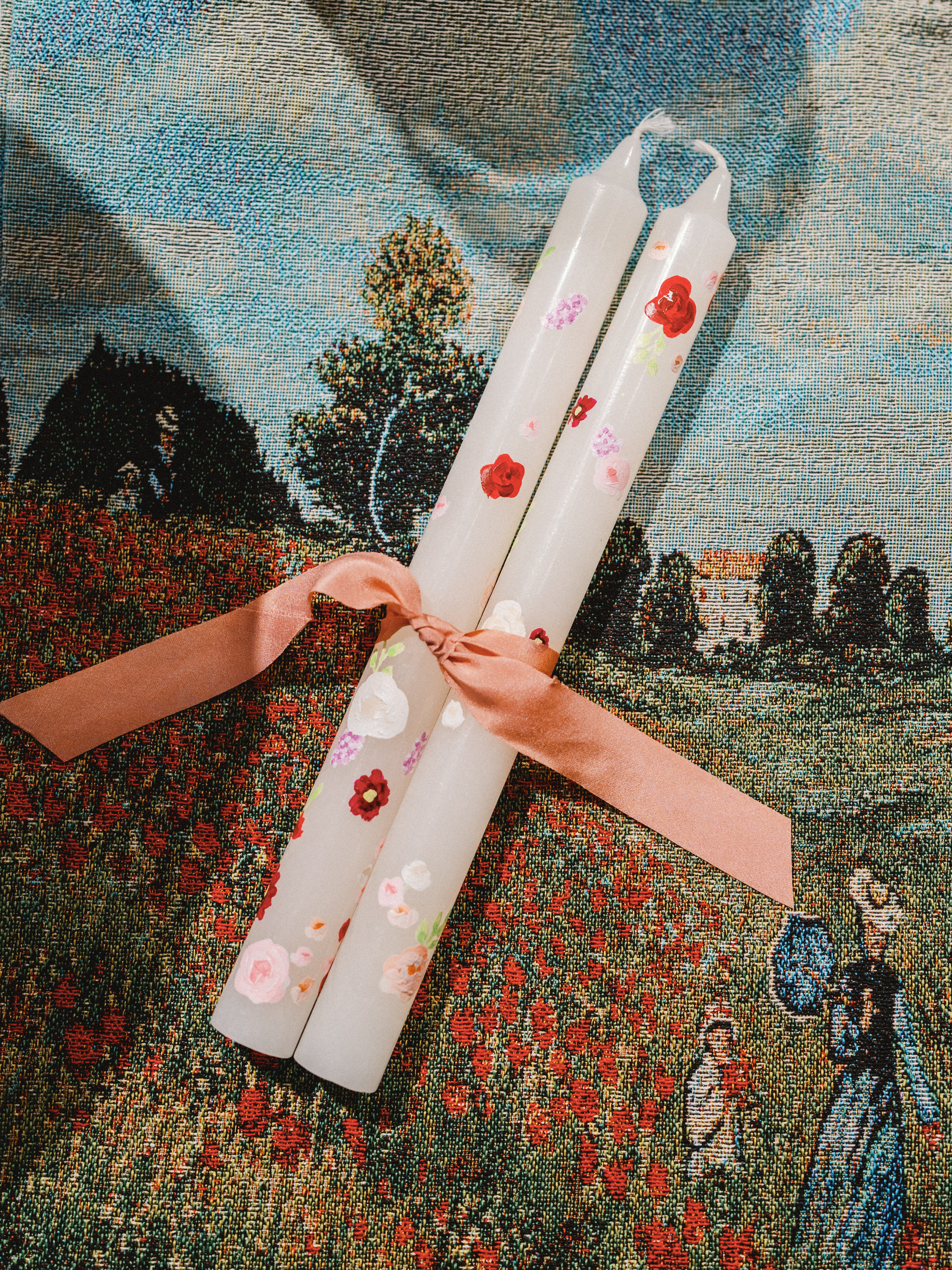 Hand-painted floral taper candles tied with a blush ribbon resting on a patterned woven textile.