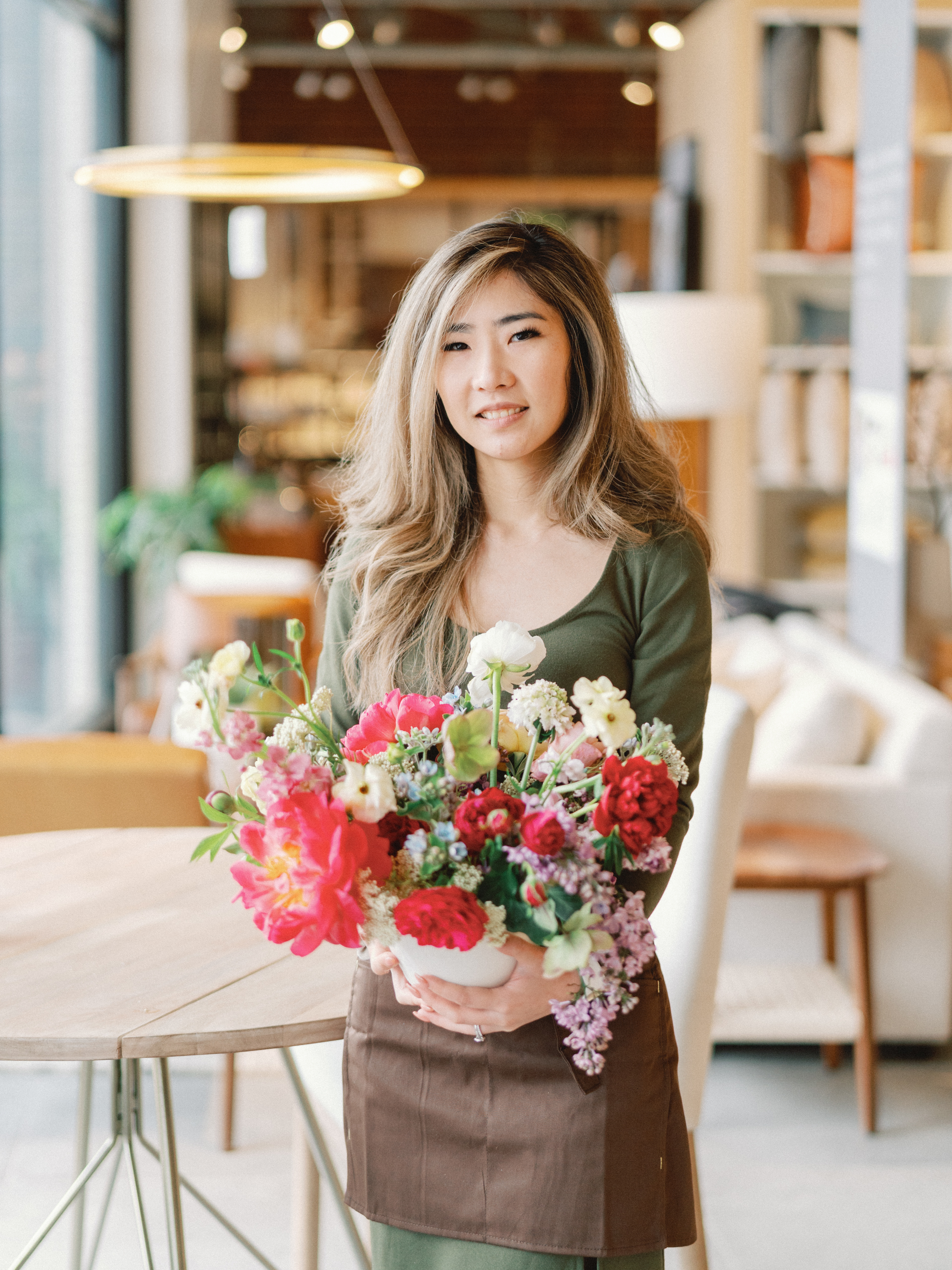 A woman smiling while holding a colorful floral arrangement featuring red, pink, and white blossoms in a white ceramic bowl.